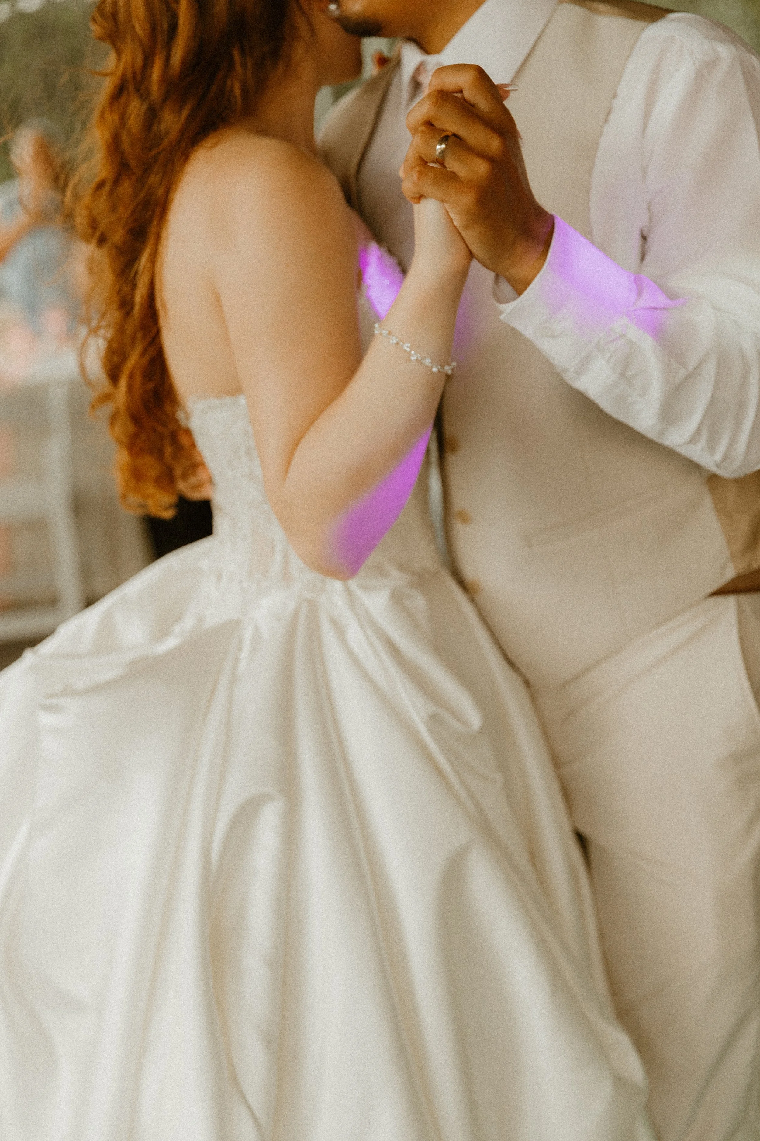 A bride and groom hold hands and dance closely at their wedding, with the bride wearing a white wedding gown and the groom in a vest and dress shirt.