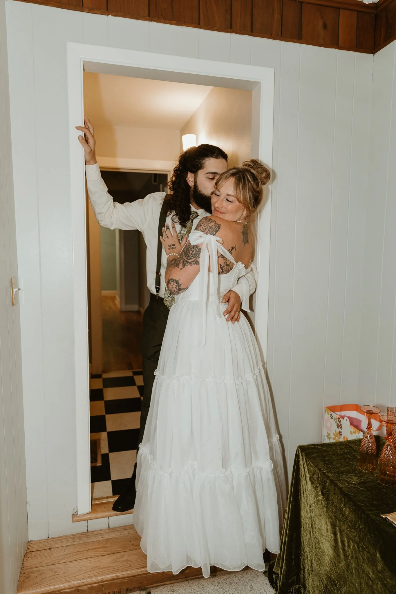 A couple sharing an intimate moment through a doorway, with the man kissing the woman on the cheek. The woman is wearing a white dress with tattoos on her arm and her hair in a bun. The man has long hair, a beard, and is dressed formally. There is a 