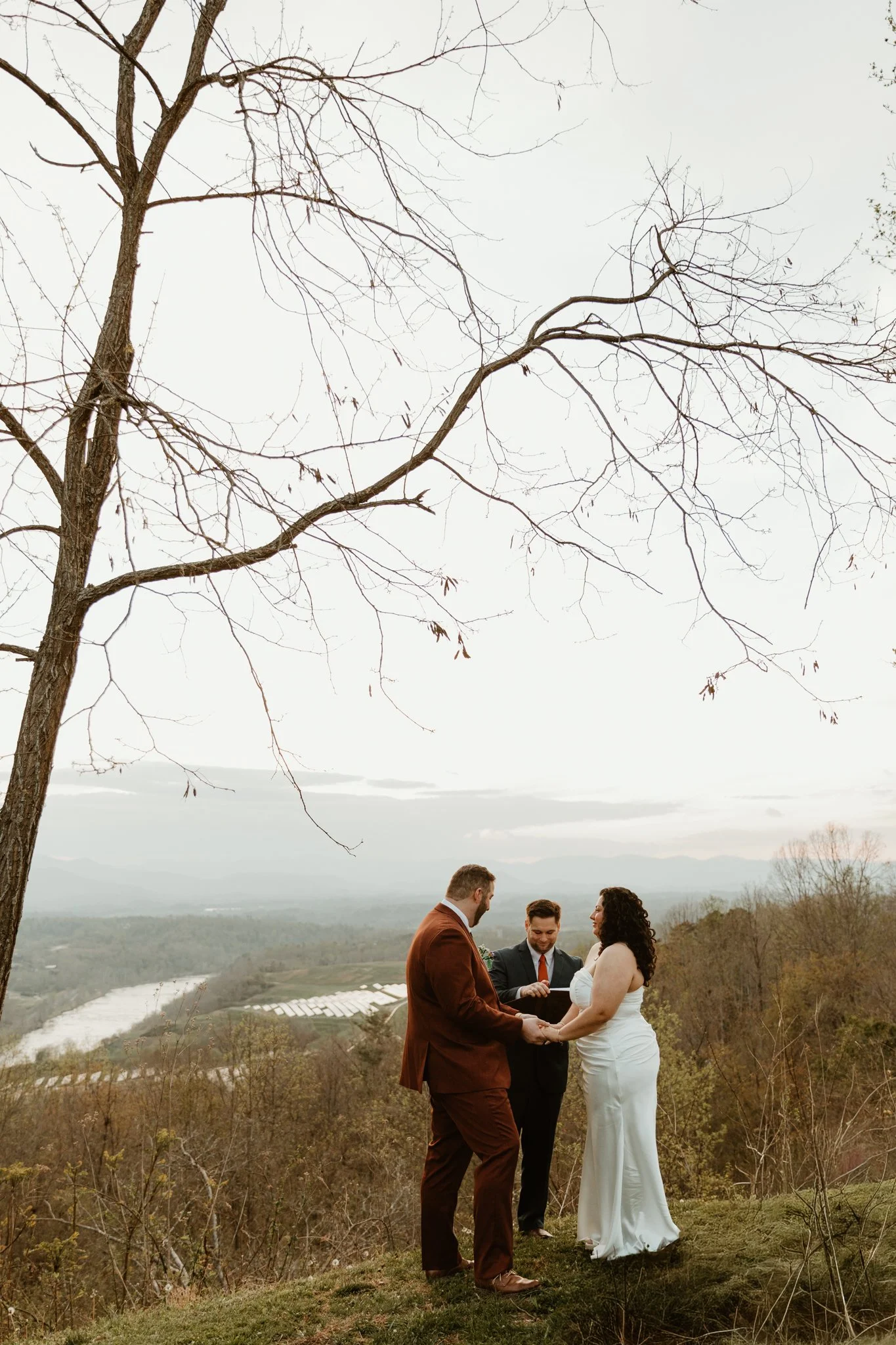 A couple and a wedding officiant holding hands during their outdoor wedding ceremony on a hill with trees and an expansive landscape in the background on a cloudy day.