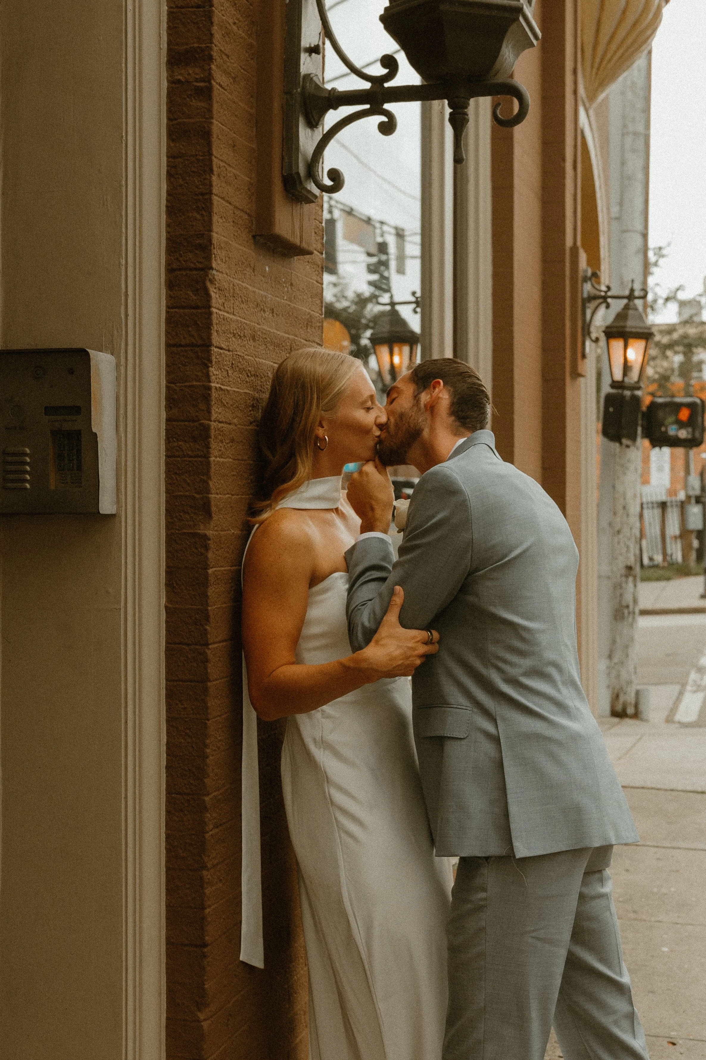 A bride and groom sharing a kiss outside a building with brick and stucco walls, lit by vintage-style wall lanterns.