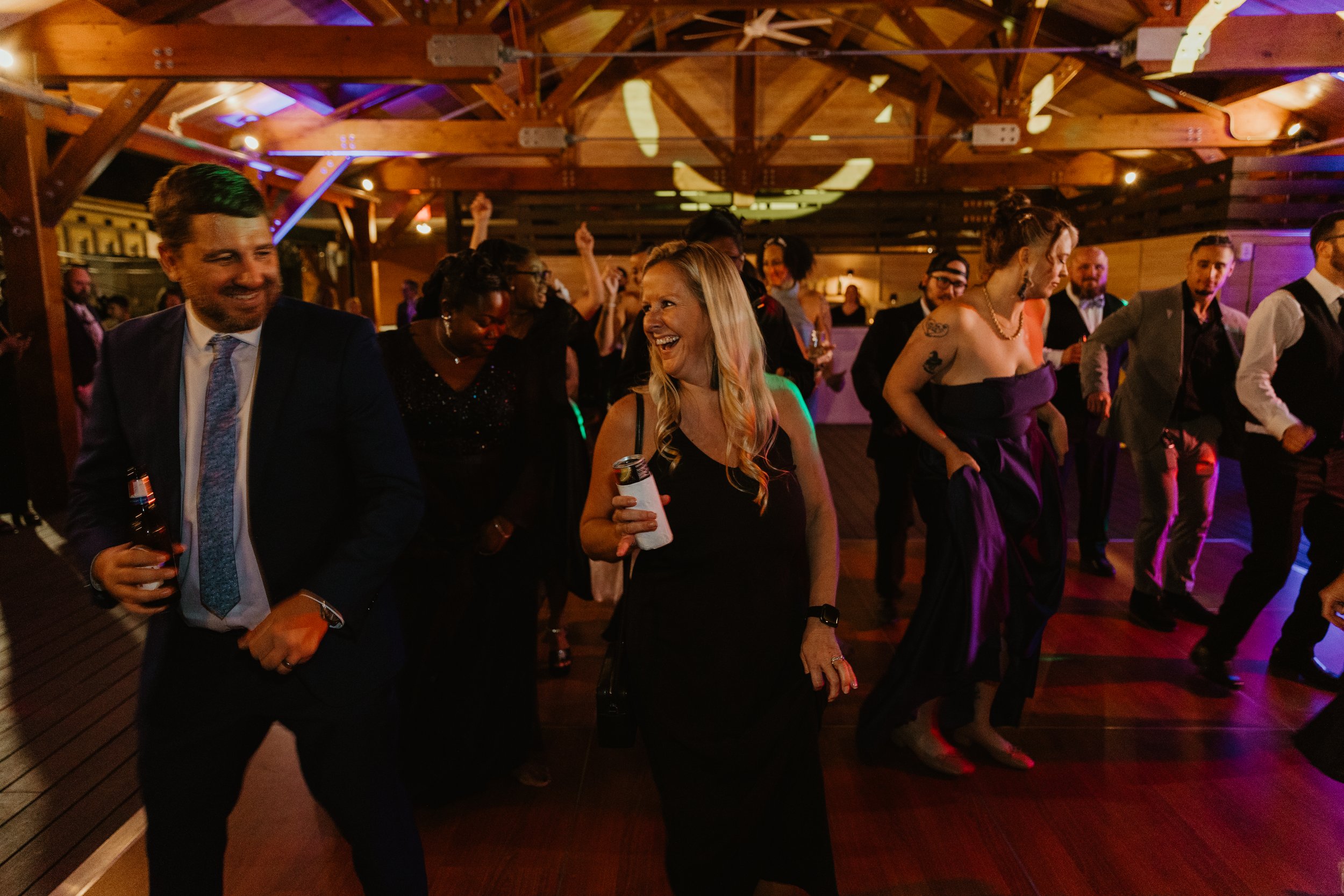 People dancing and socializing at a party or celebration indoors with wooden beams and colorful lighting.