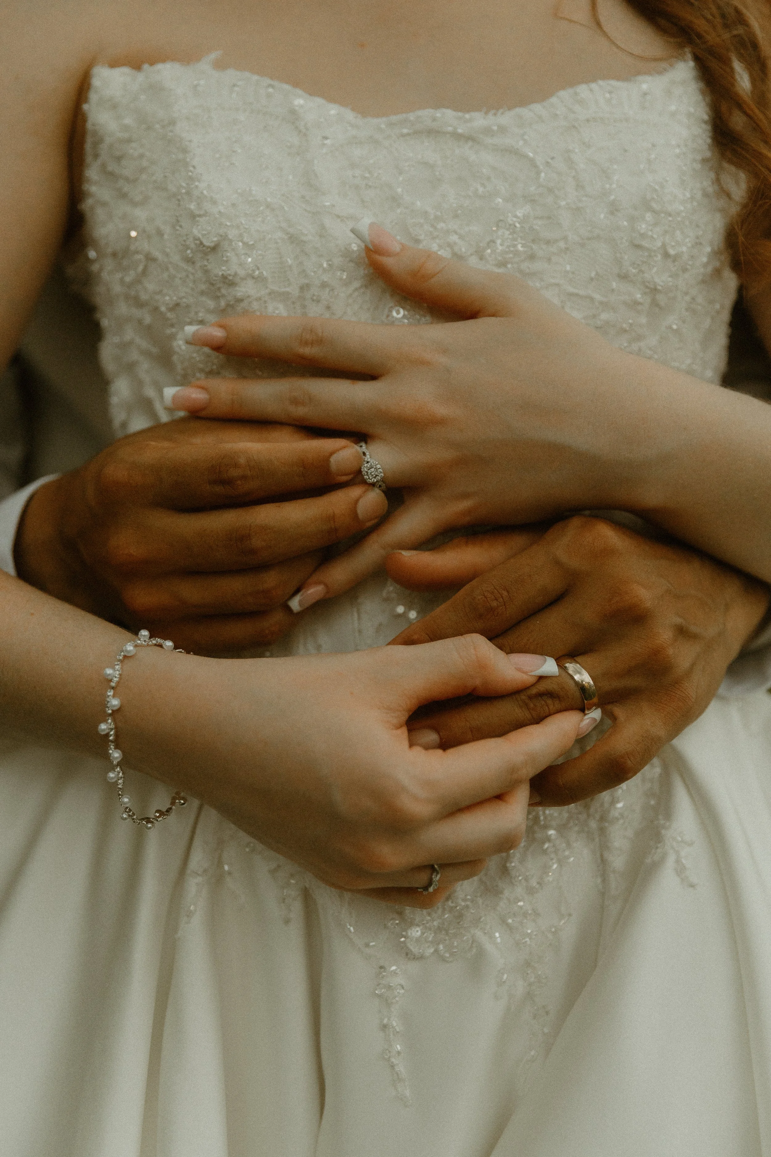 Close-up of a bride's hands with a wedding ring, resting on her wedding dress. Several other hands are gently holding her hands, symbolizing support and love.
