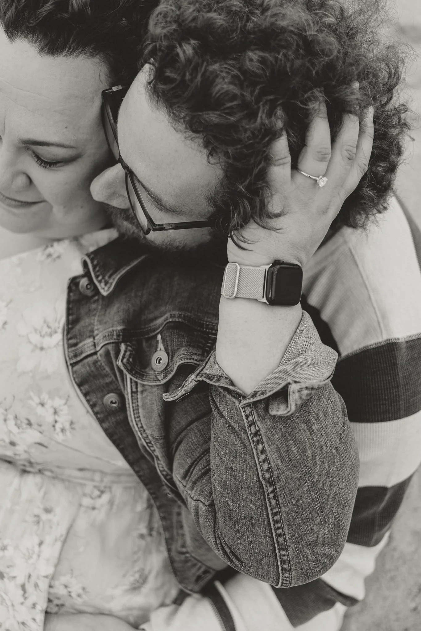 A close-up of a couple in an intimate embrace, with the man holding the woman's face and leaning his forehead against hers. The woman is wearing glasses and a denim jacket, and the man is wearing a smartwatch and has a ring on his finger. They are ou
