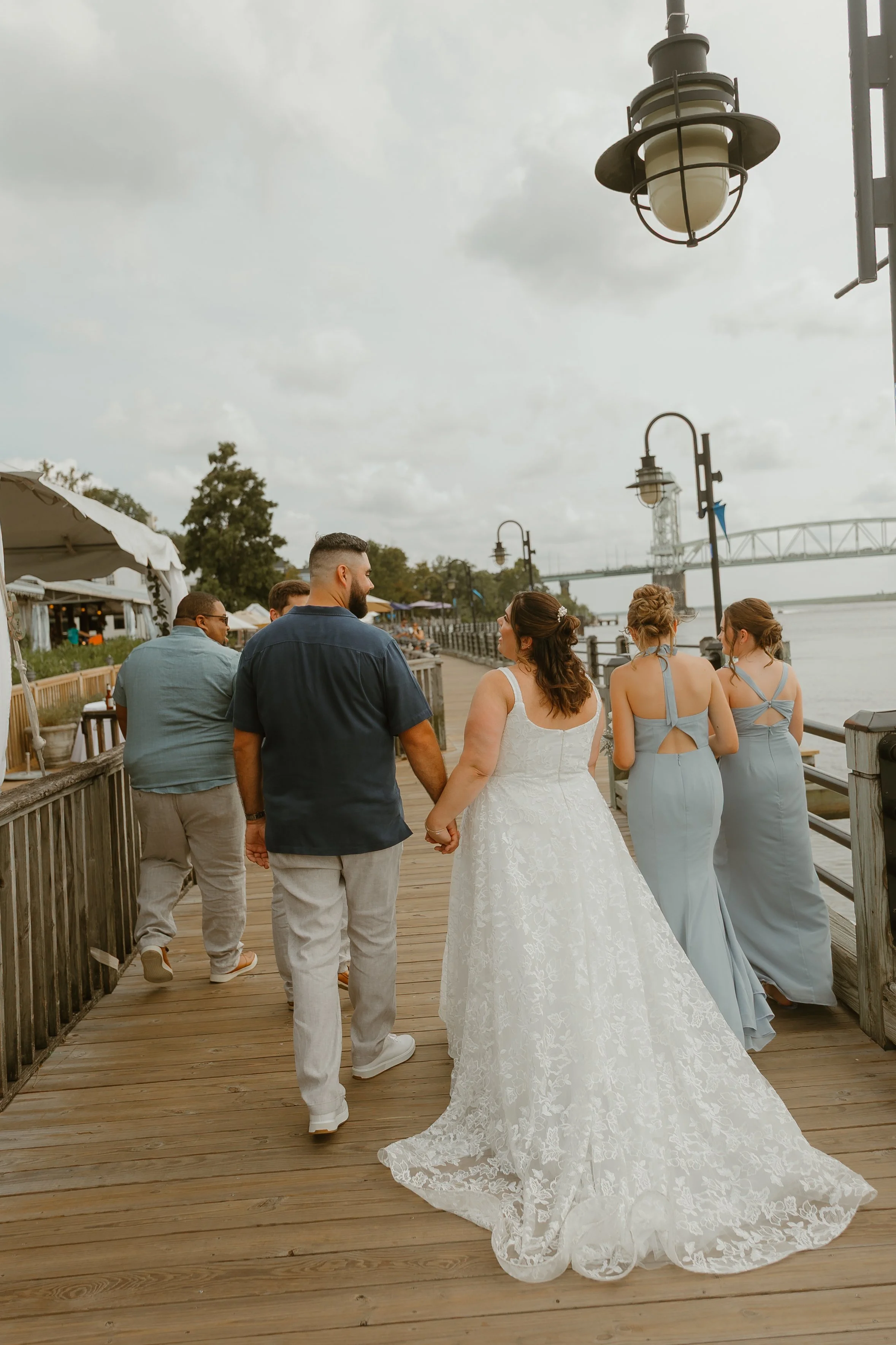 A bride in a white lace wedding dress and her groom holding hands walk on a wooden boardwalk with friends nearby. The bride’s hair is styled in loose waves with a floral accessory. The groom is wearing a navy shirt and light-colored pants. Other wome