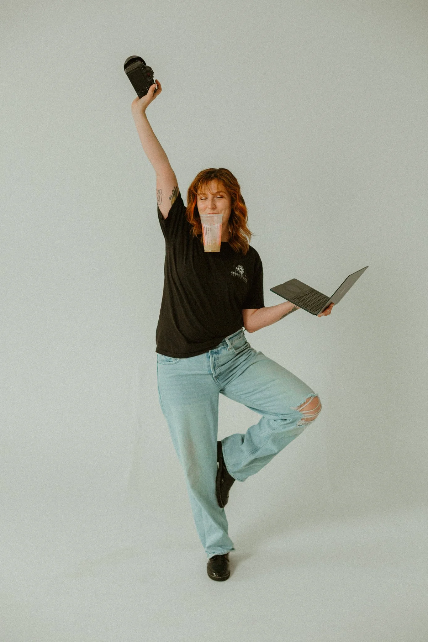 A woman with red hair wearing a black T-shirt, ripped jeans, and black shoes, balancing on one leg with her other foot raised, while holding a small laptop in one hand, a camera in her extended other hand, and a snack package near her mouth, against a plain gray background.
