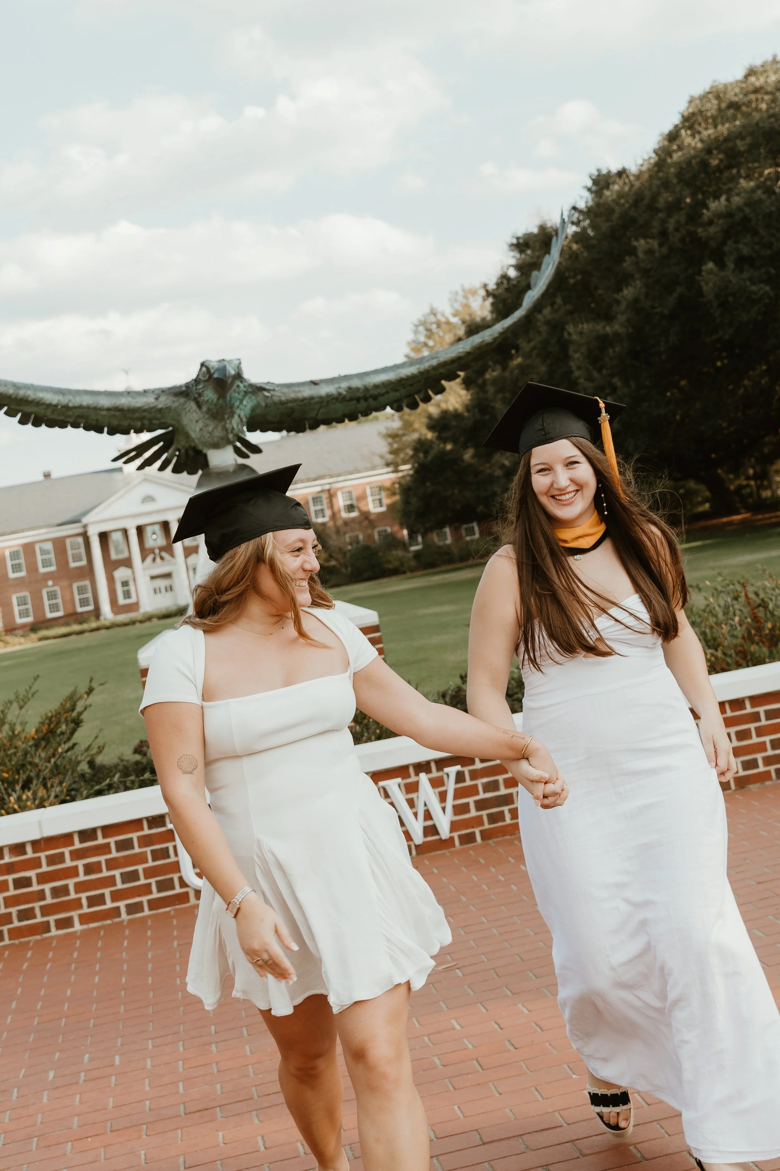 A young woman in graduation cap and gown standing in front of a fountain with columns during autumn.