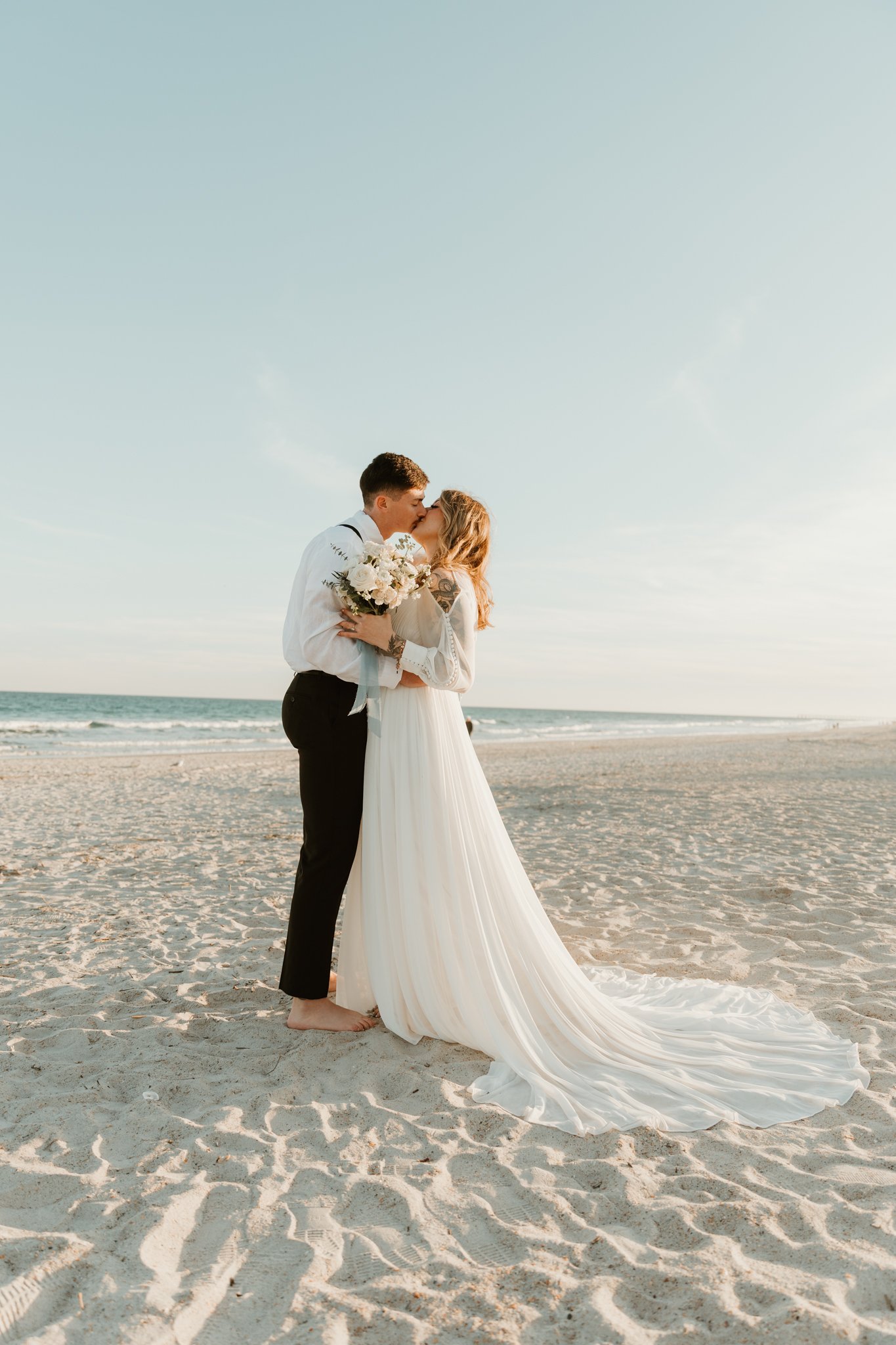 A couple in wedding attire sharing a kiss on a beach at sunset, with the bride holding a bouquet of flowers.