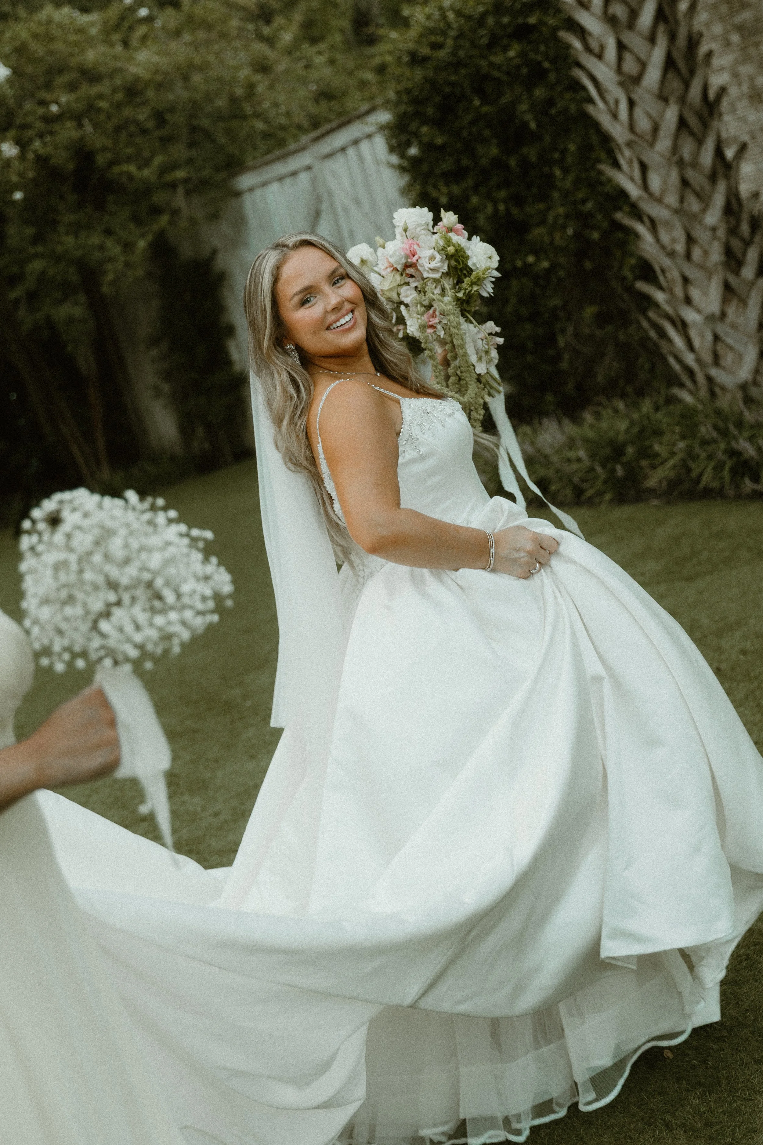 A smiling bride in a wedding dress holding a bouquet of pink and white flowers outdoors.