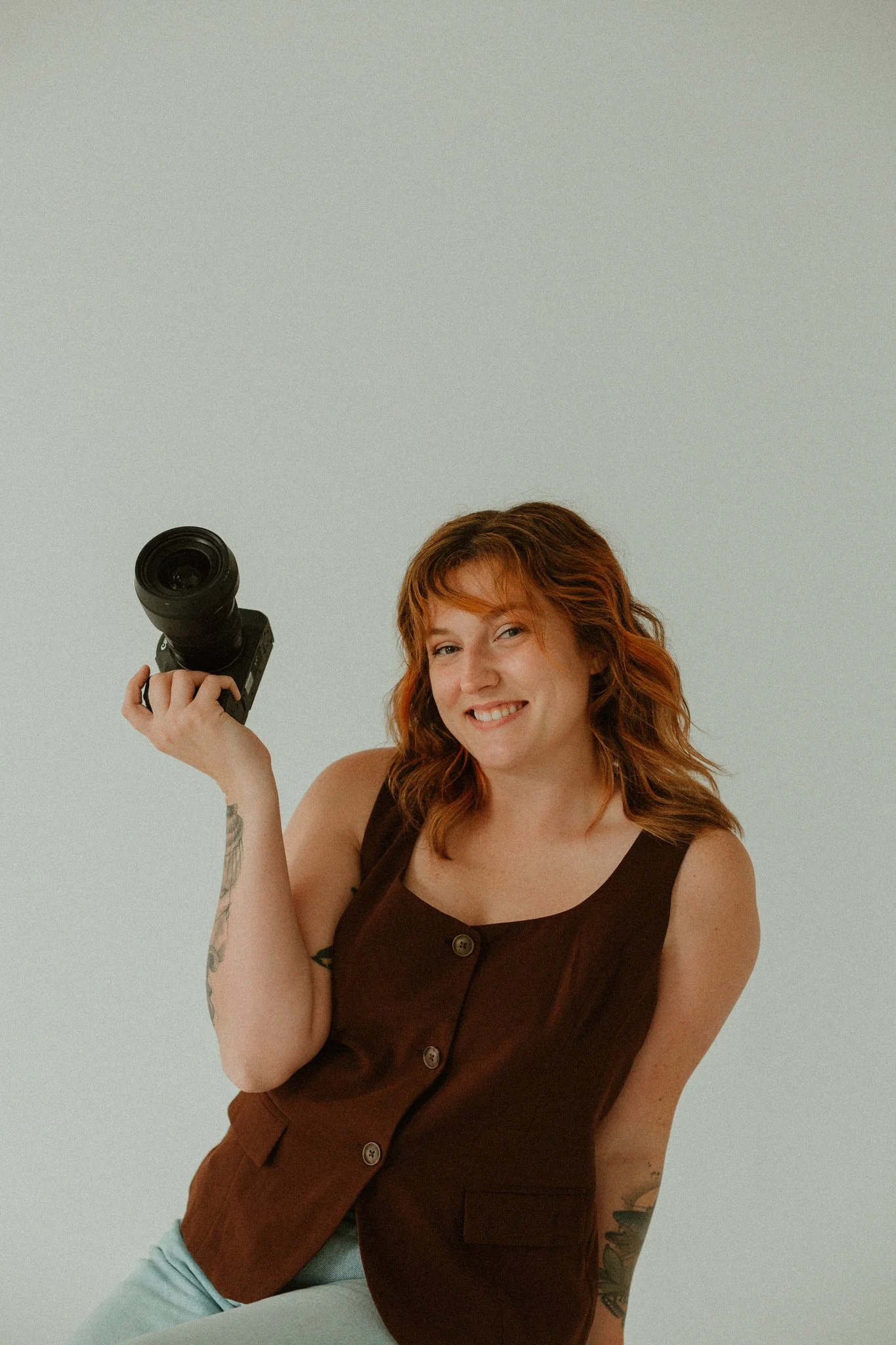 A woman with red hair smiling, holding a camera, wearing a brown sleeveless top, seated against a plain light-colored background.