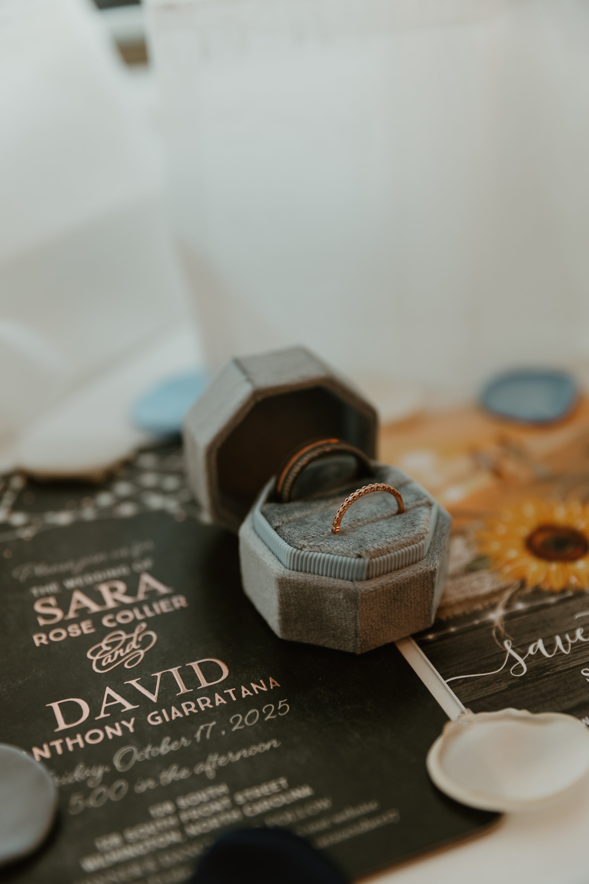 A wedding invitation with an open ring box containing a gold ring and a wedding band placed on top. The invitation features text including the names Sara and David, and the date October 11, 2025. The scene is decorated with invitations and floral ele