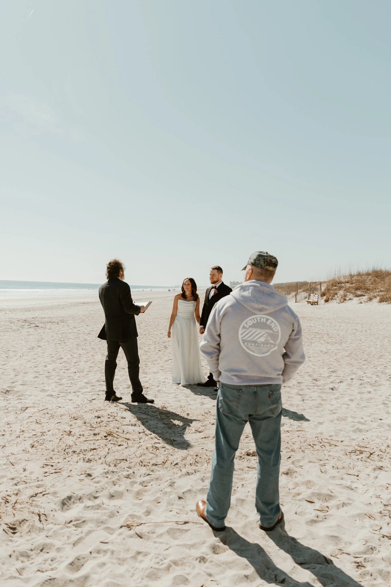 A wedding ceremony taking place on a sandy beach with a bride in a white dress and a groom in a suit holding hands, officiant speaking, and a man in a hoodie and baseball cap watching.