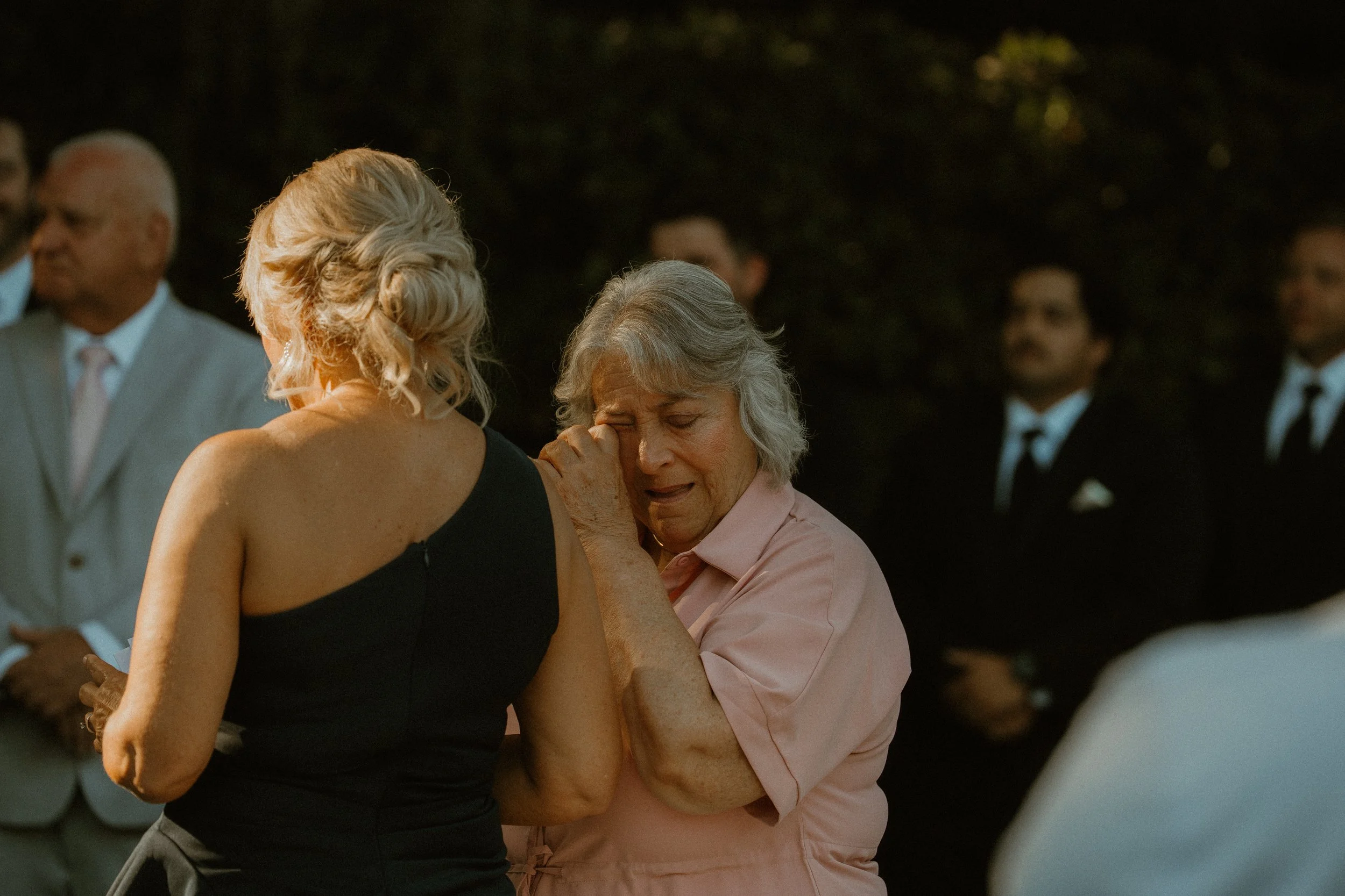An elderly woman with gray hair and a pink shirt hides her face in her hands and appears emotional, while a woman with blonde hair in a black dress comforts her. Several people in formal attire stand nearby in an outdoor setting at dusk.