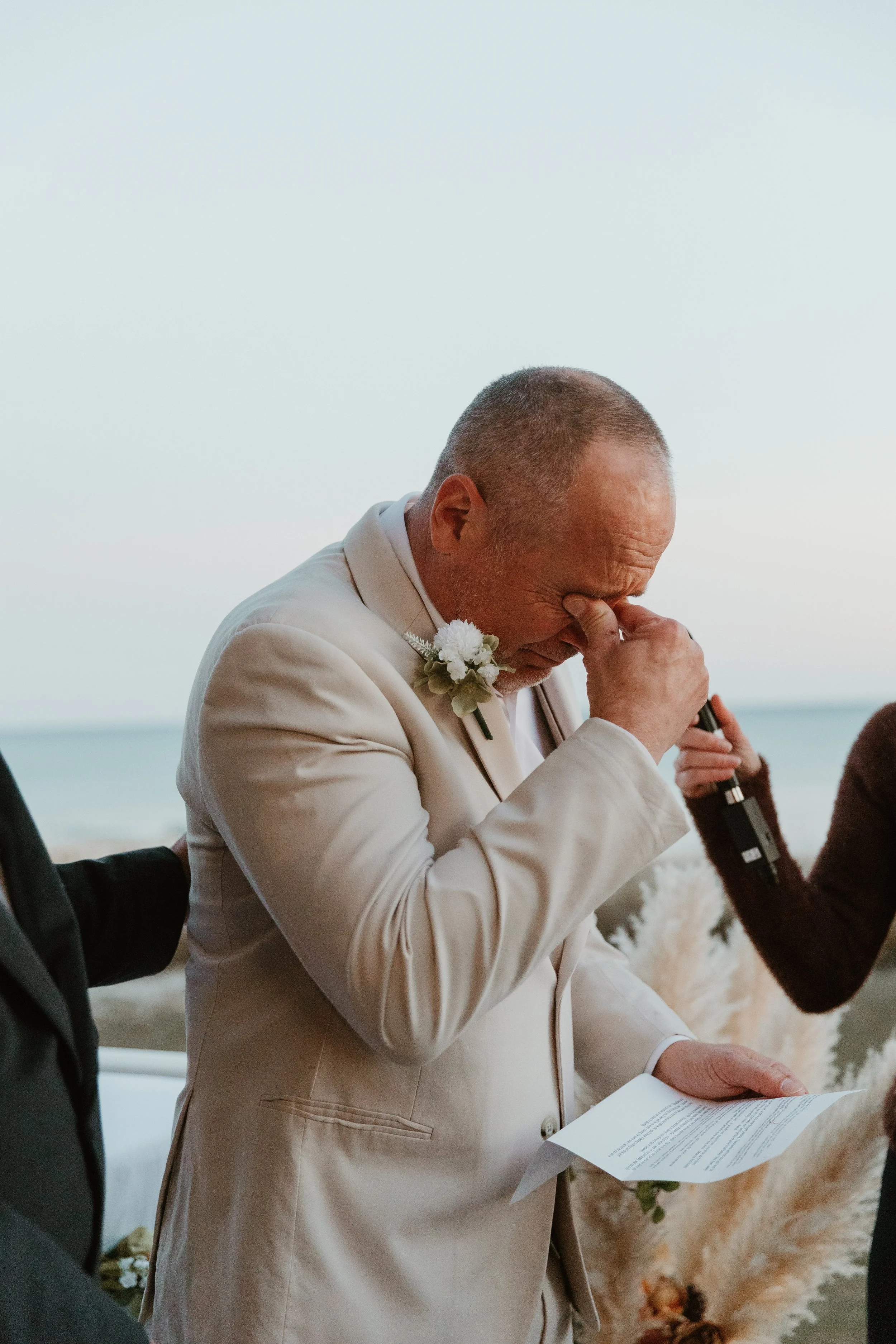 An older man in a beige suit and white shirt is emotionally crying during a wedding ceremony on the beach, holding a piece of paper.