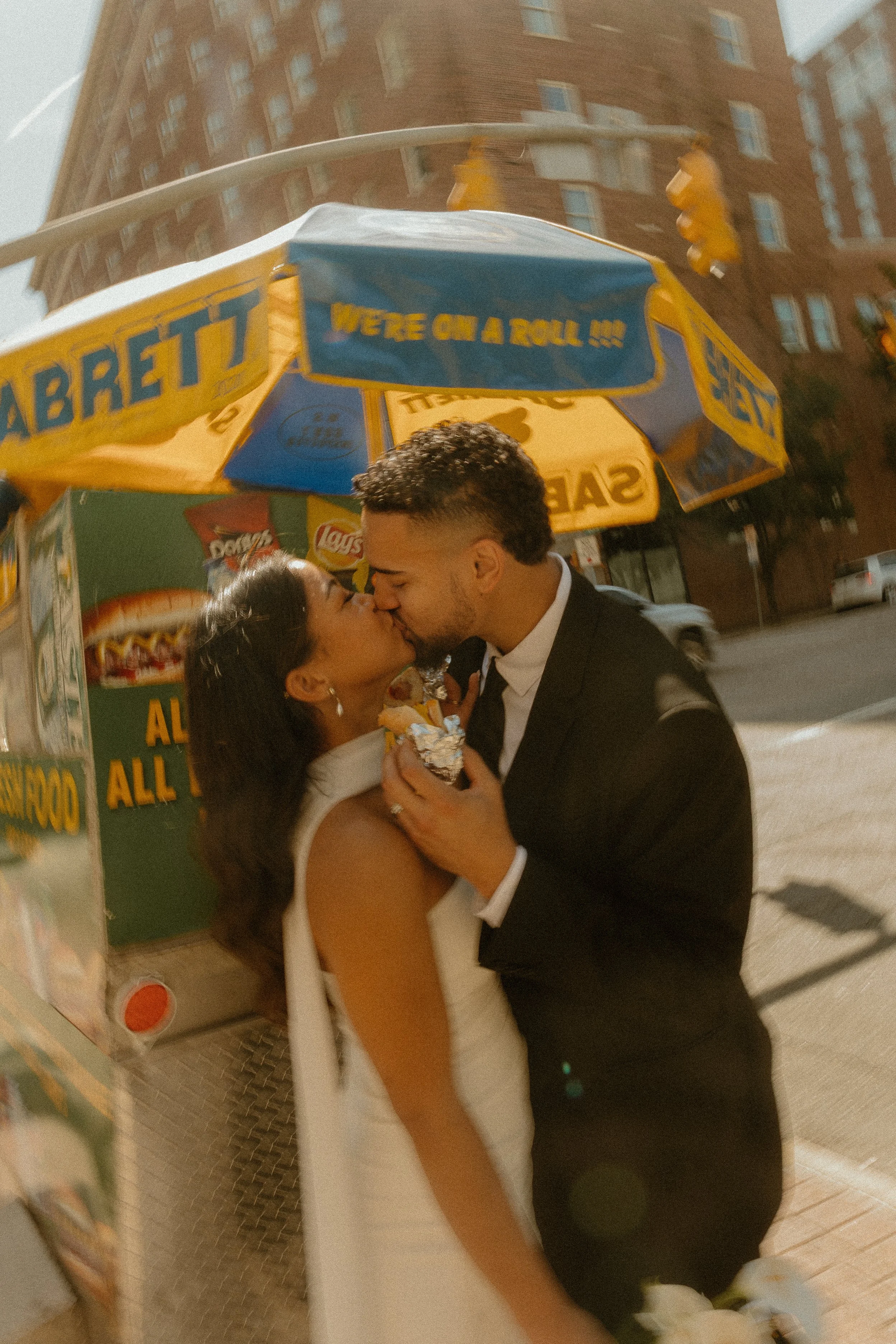 A couple in formal attire sharing a kiss in front of a food cart on a city street.