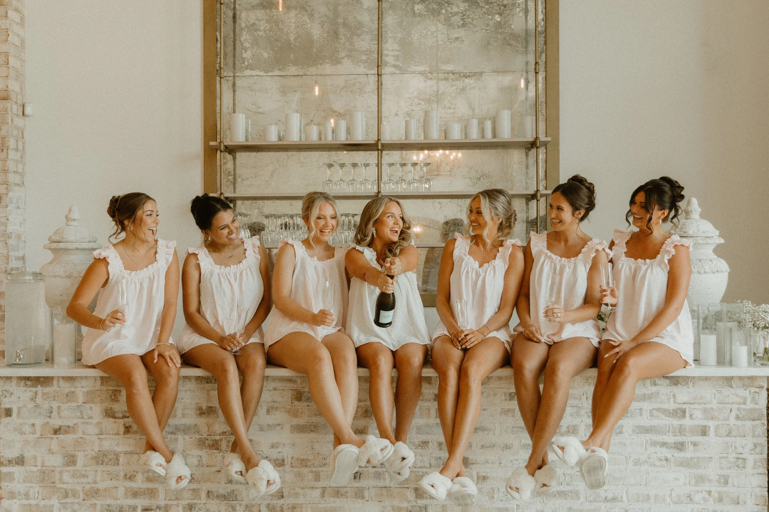 A group of seven women sitting on a brick ledge, wearing matching white dresses and slippers, smiling and laughing at a celebration, with candles and glasses on a shelf behind them.