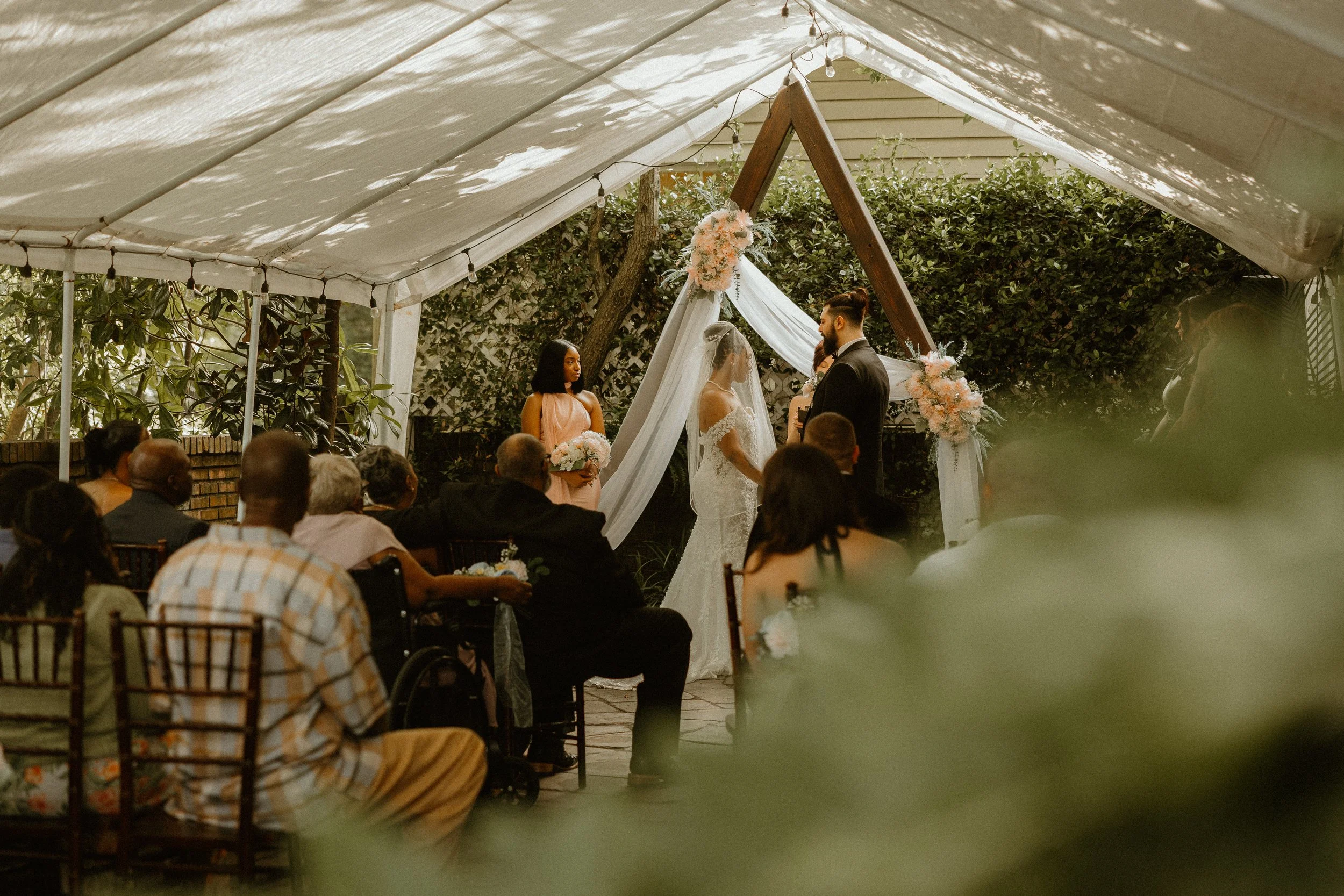 A wedding ceremony taking place outdoors under a white tent with a floral arch. The bride and groom stand face to face, surrounded by seated guests. The scene is decorated with pink and white flowers and greenery.