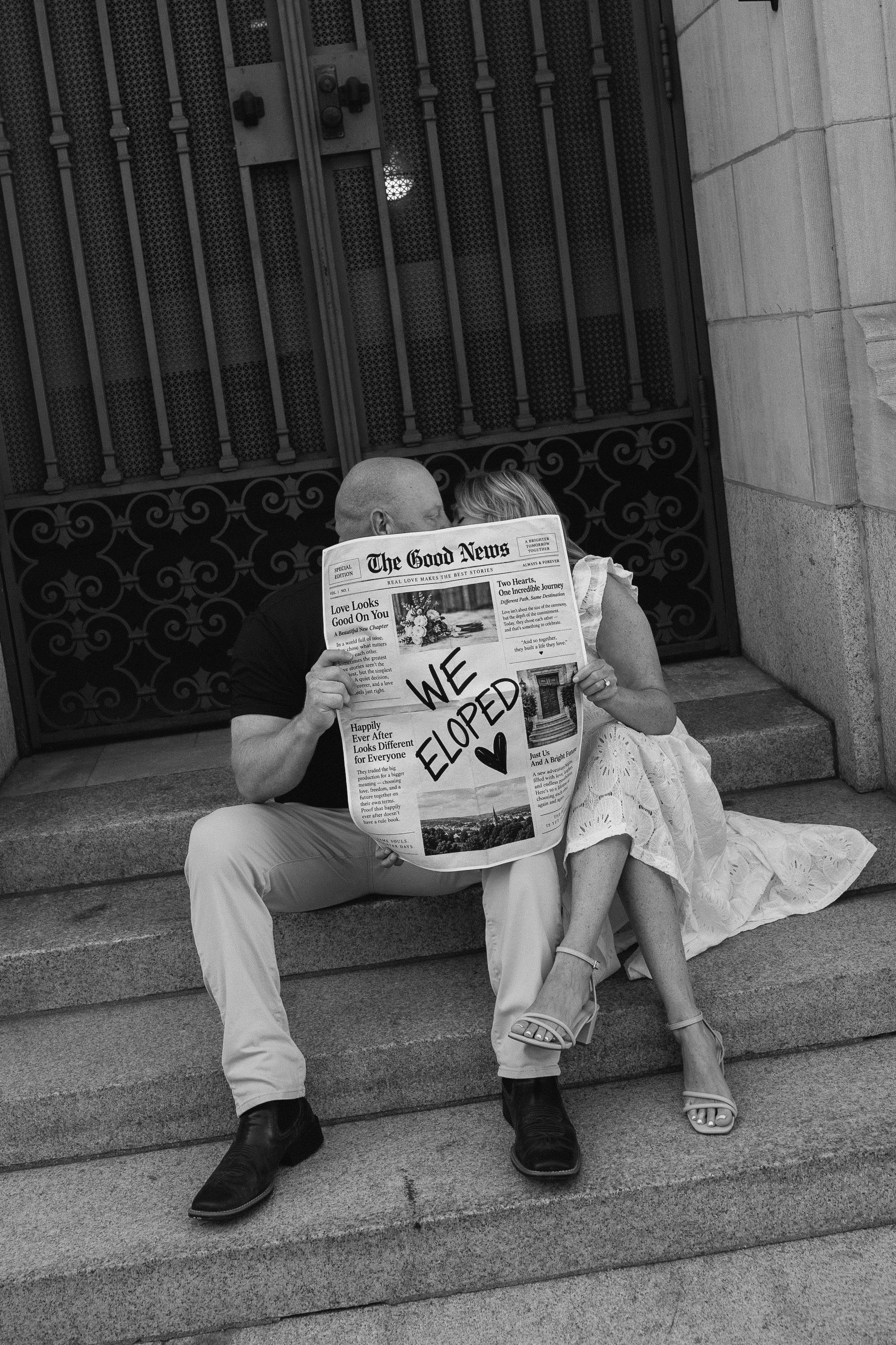 A couple is sitting on steps outside a building, reading a newspaper that has a handwritten message saying, 'WE '. The man is dressed in a dark shirt and light pants, and the woman is wearing a light dress and sandals. The background features a decor