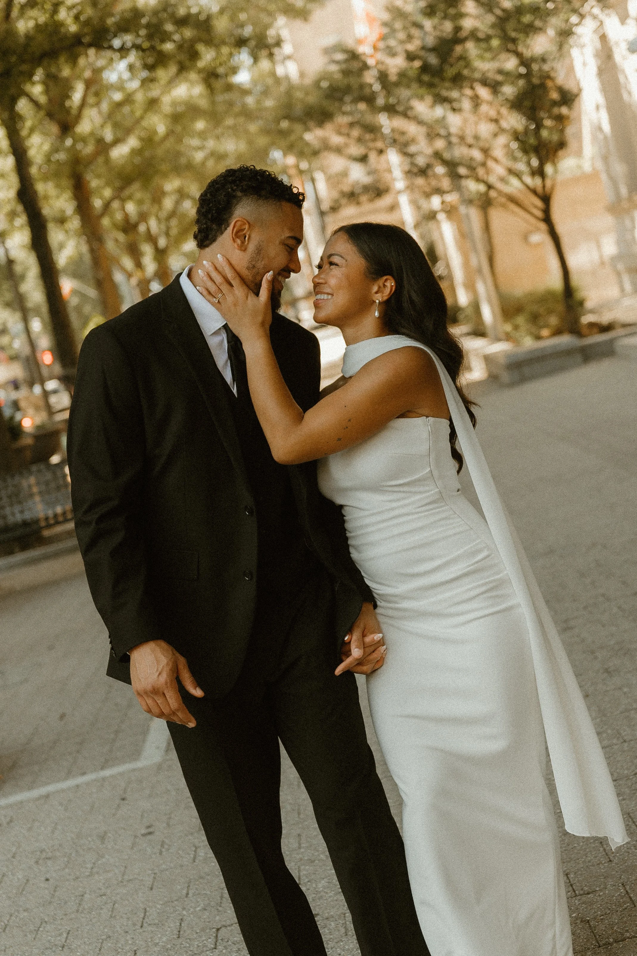 A joyful couple, dressed in formal attire, stands close together outdoors under the trees, gazing into each other's eyes and holding hands, sharing a tender moment.
