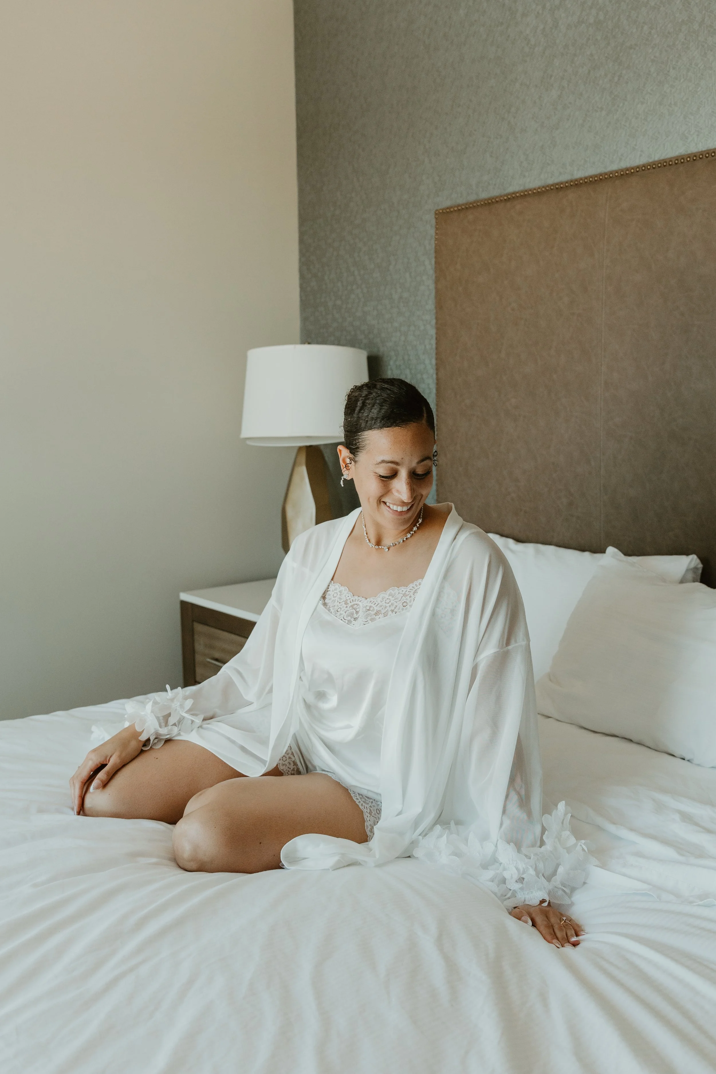 A smiling woman in white satin and lace lingerie and a robe, sitting on a bed in a hotel room, looking down.