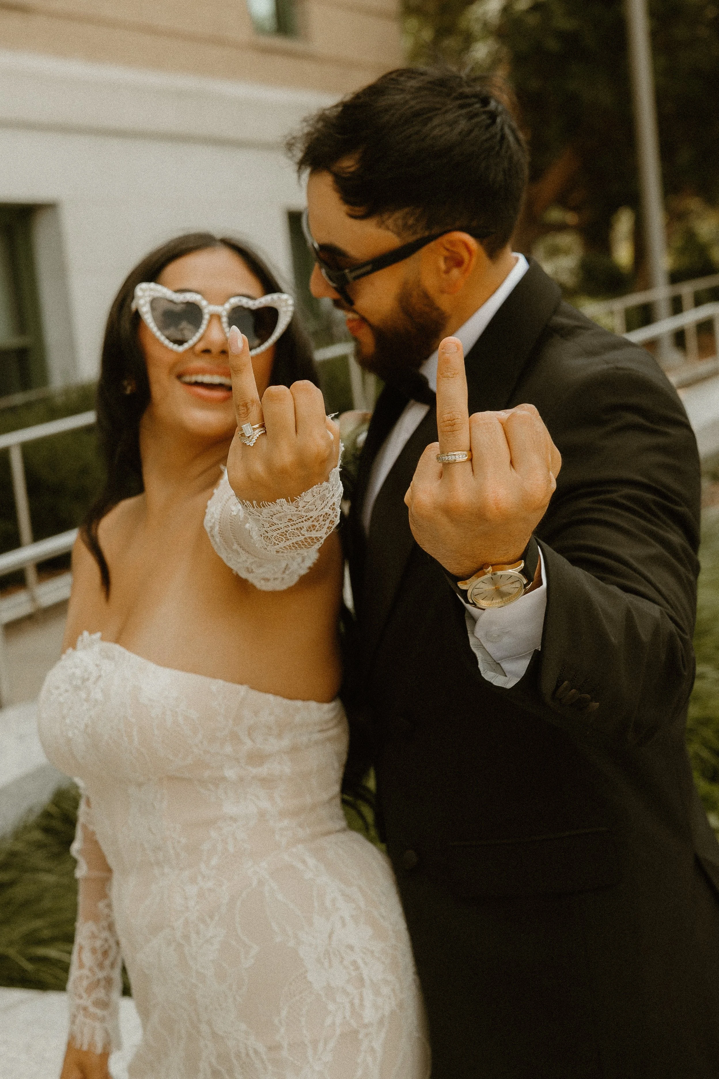 A newly married couple is celebrating and showing excitement, with the bride wearing heart-shaped sunglasses and the groom wearing glasses, both displaying their middle fingers.