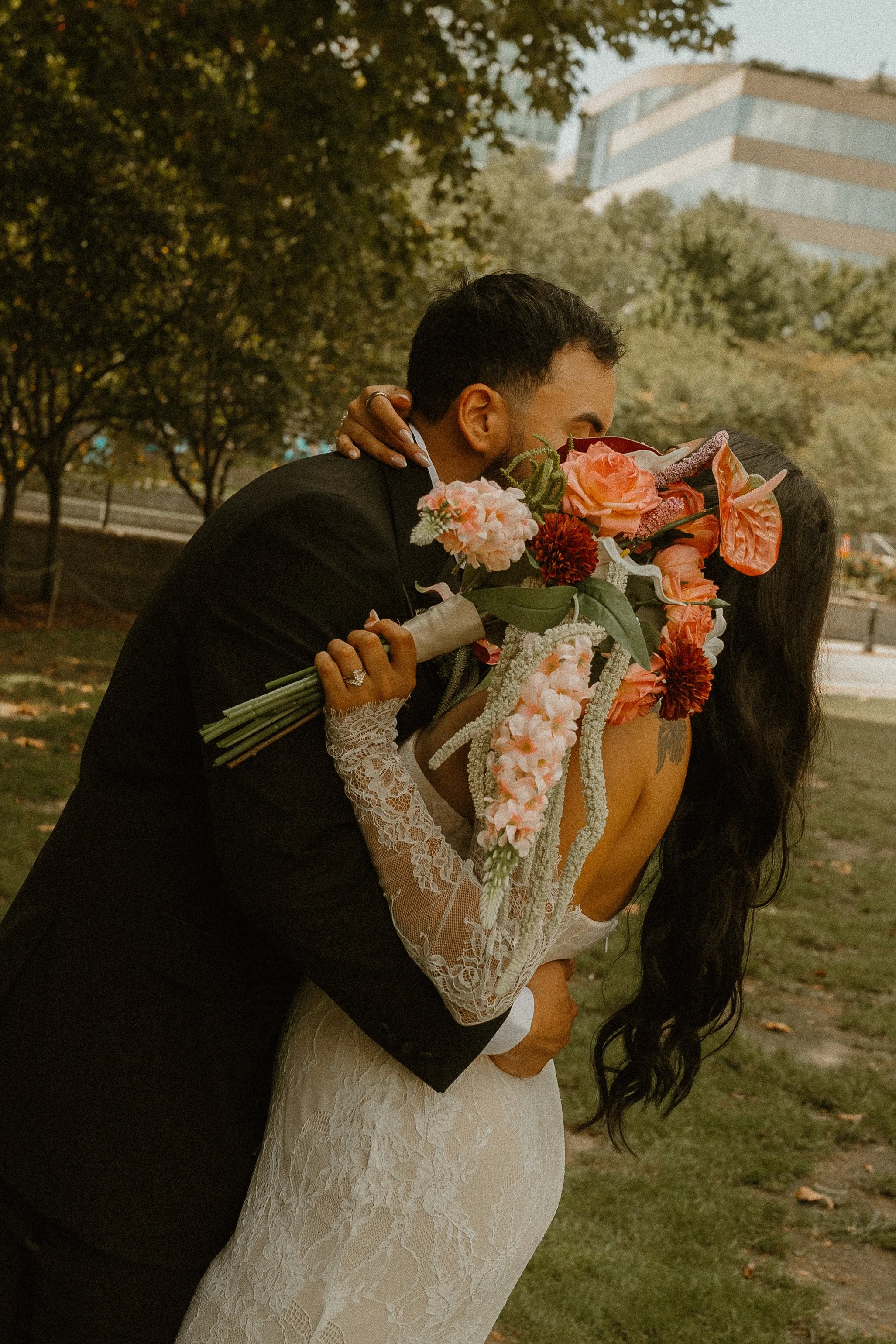 Couple kissing outdoors, woman holding a large bouquet of flowers, woman in lace dress, man in dark suit, trees and modern building in background.