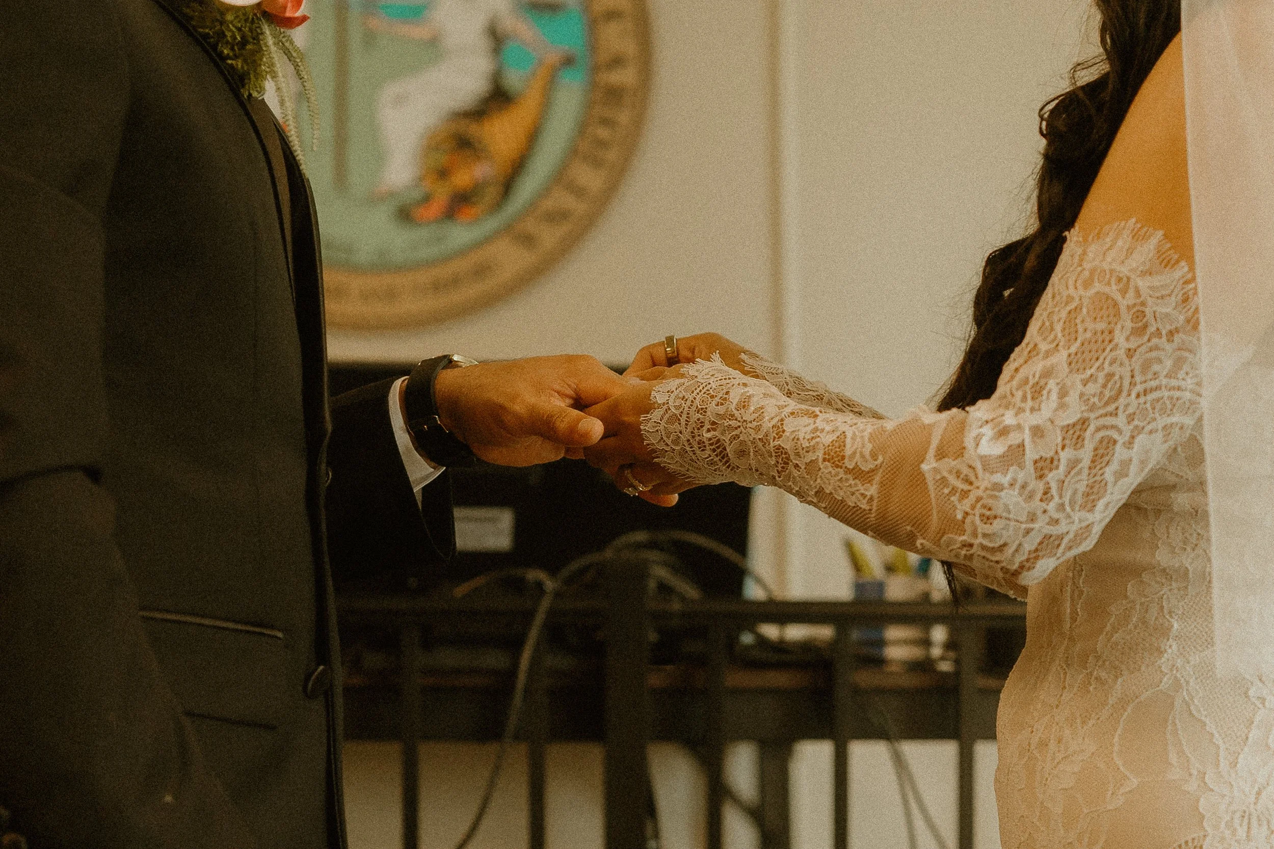A woman with long dark hair wearing a lace cream-colored dress is holding hands with a man dressed in a dark suit, in an indoor setting with a wall and a colorful painting in the background inside the Buncombe County Courthouse