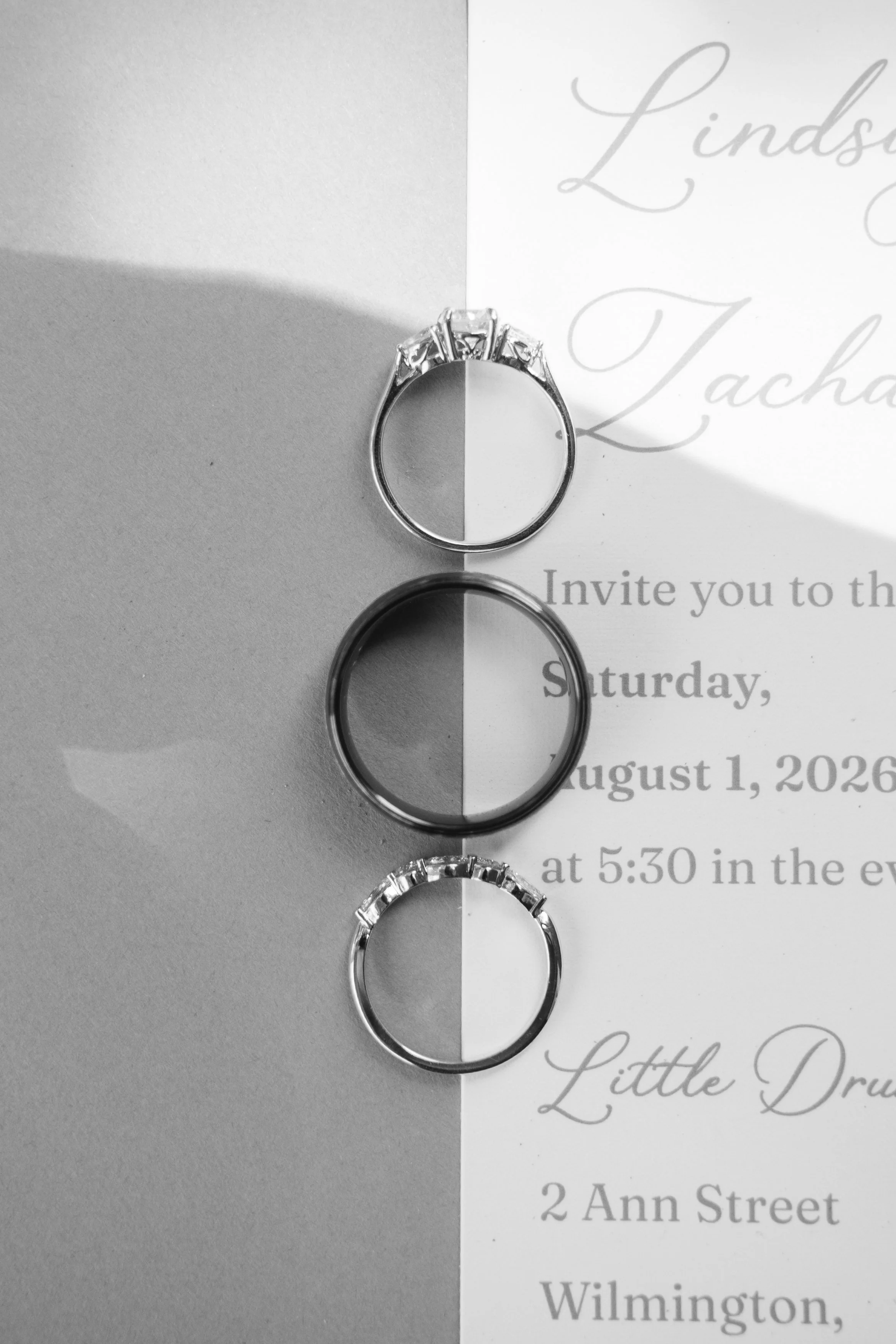 Black and white photo of three rings placed on a wedding invitation card. The rings are aligned vertically, with the top ring being a special engagement ring with side stones, the middle ring is a plain band, and the bottom ring has a design with mul