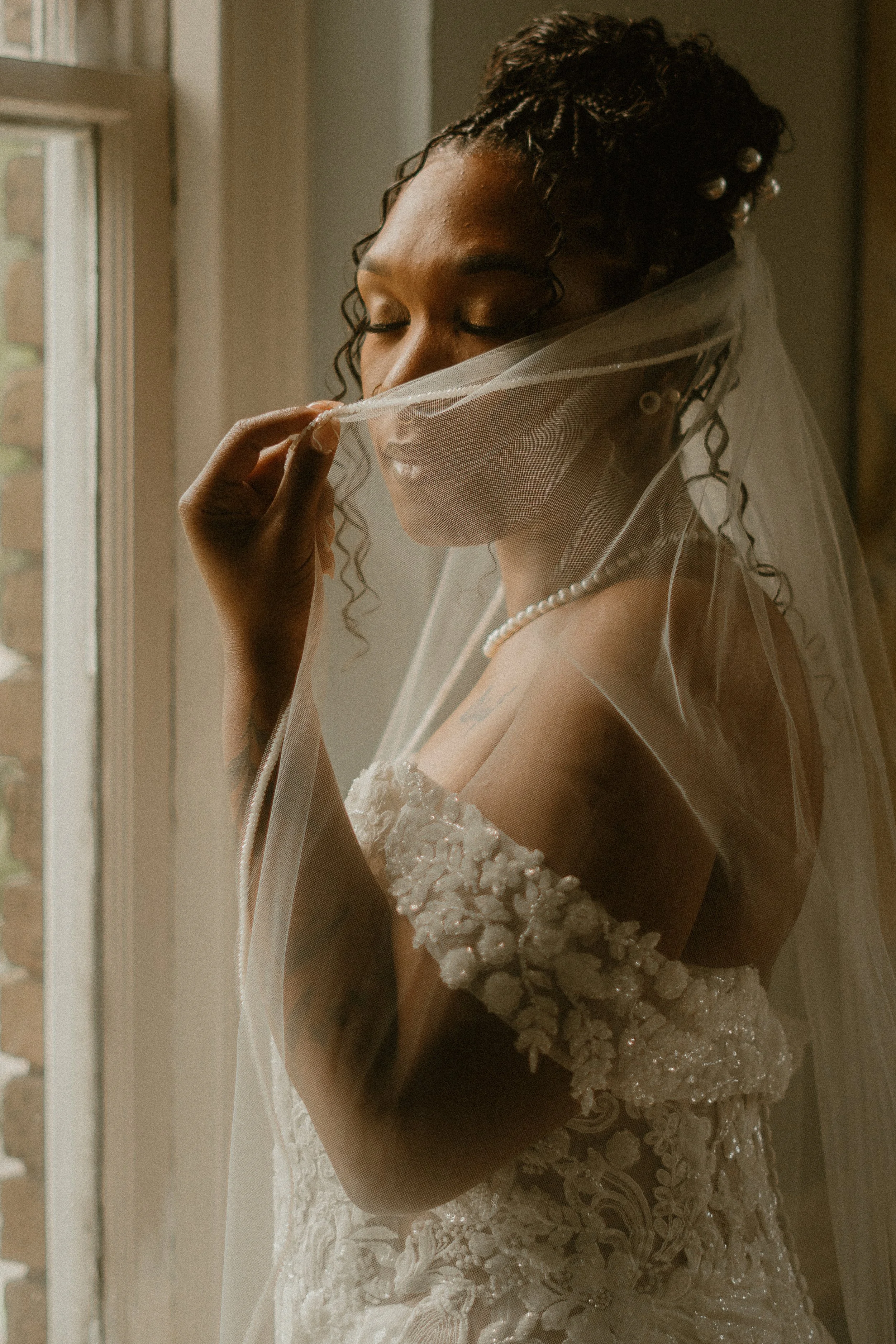 A bride with closed eyes, holding a sheer veil to her face, wearing a detailed lace wedding dress with floral appliqué and pearls, standing near a window with soft natural light