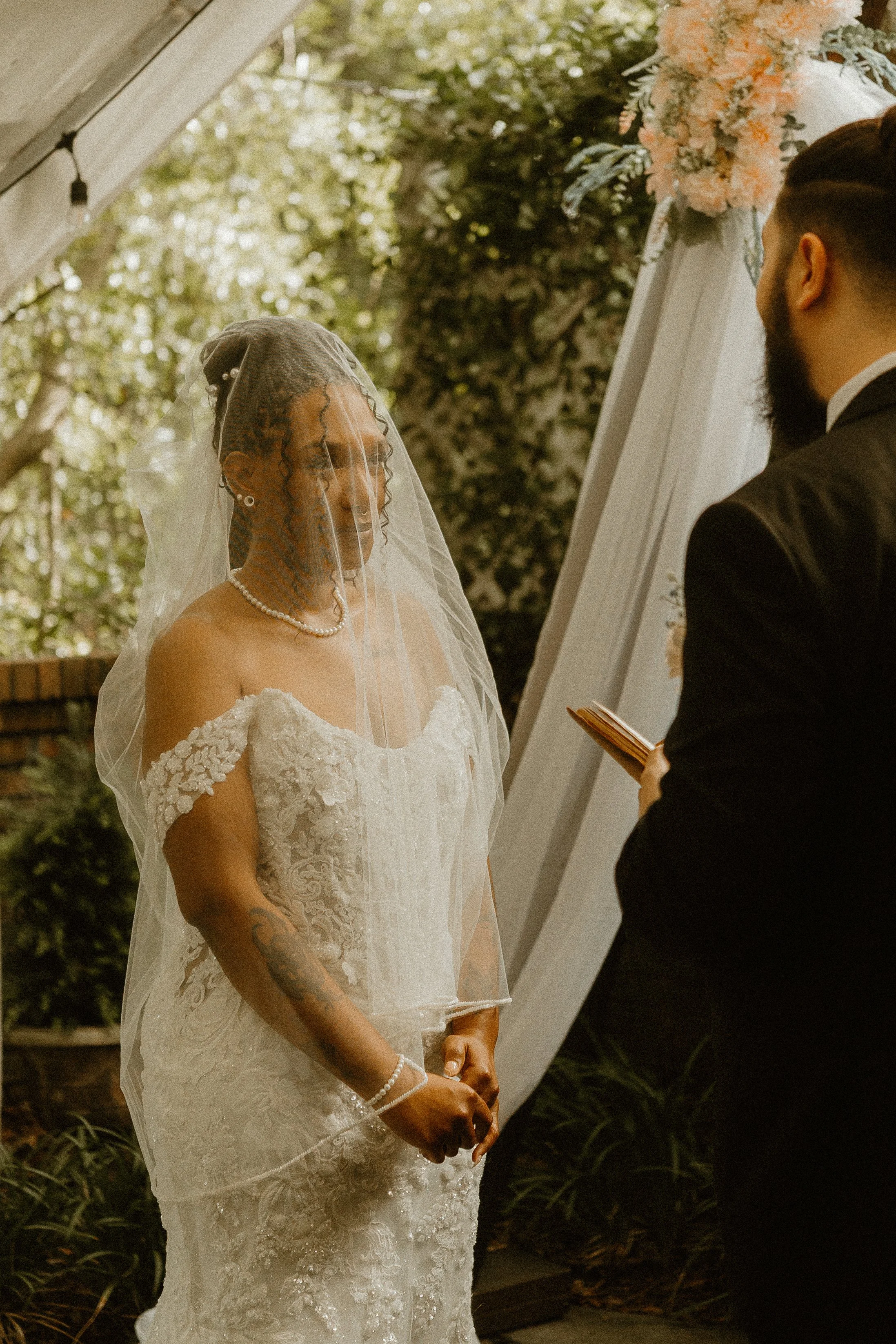 A bride and groom exchanging vows during an outdoor wedding ceremony with a floral arch and greenery in the background.