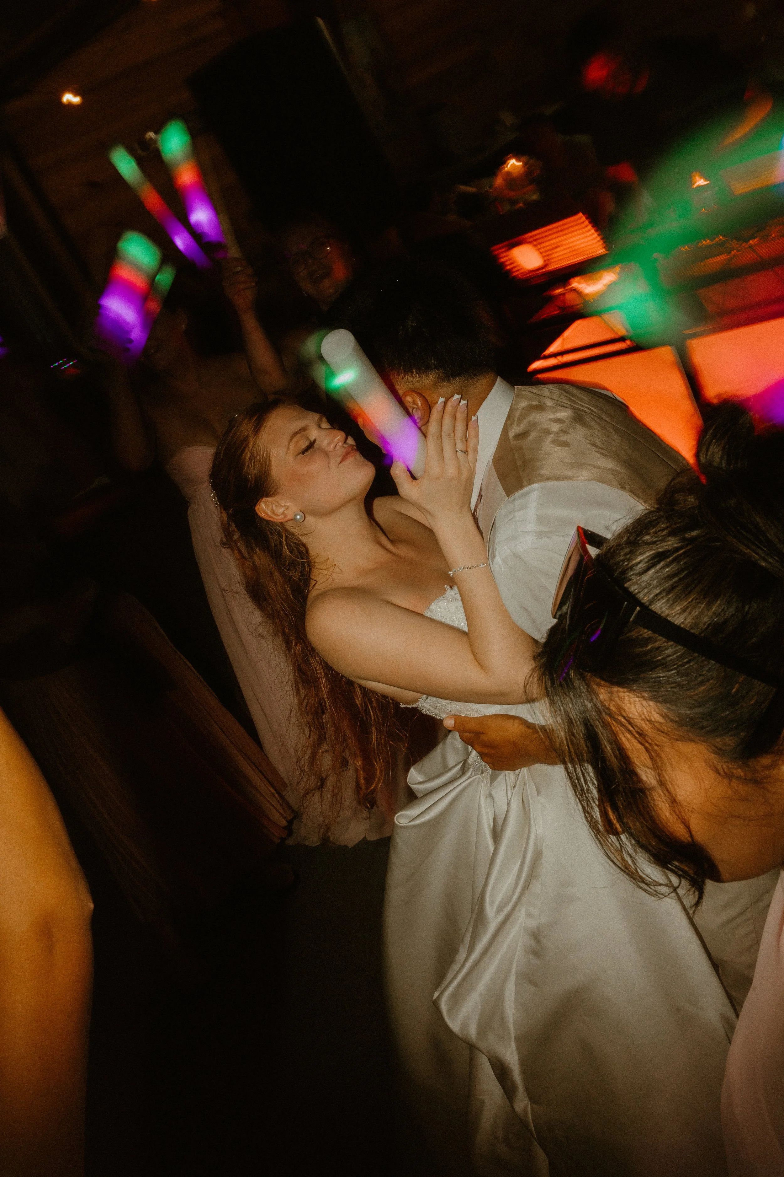 Woman in white dress dancing with man in vest in dimly lit venue with colorful lights.