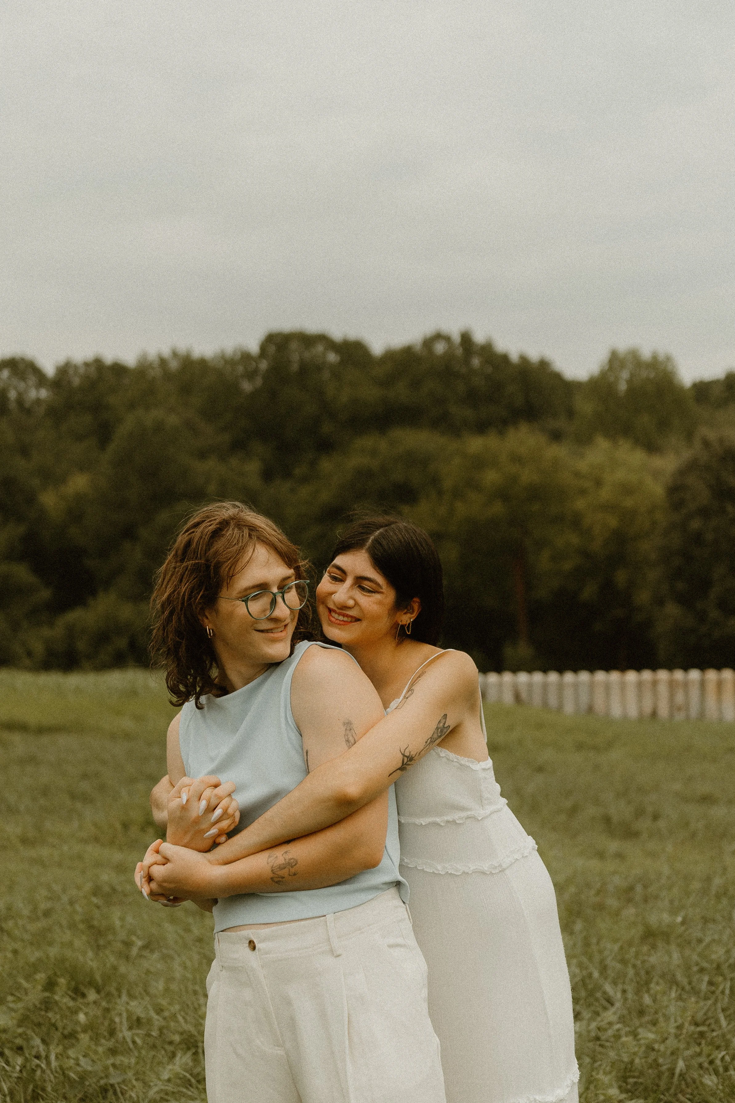 Two women embracing outdoors on a grassy field with trees in the background, smiling and looking happy.