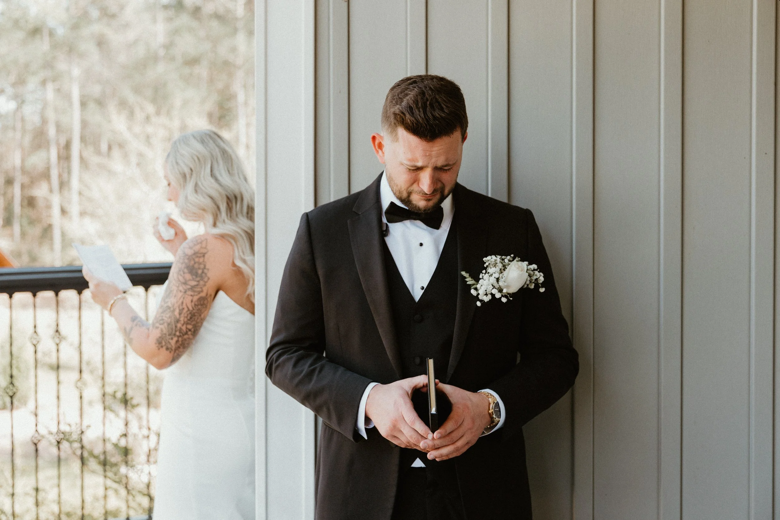 A groom dressed in a black tuxedo with a bow tie, holding a book, standing against a gray wall with a woman in the background.