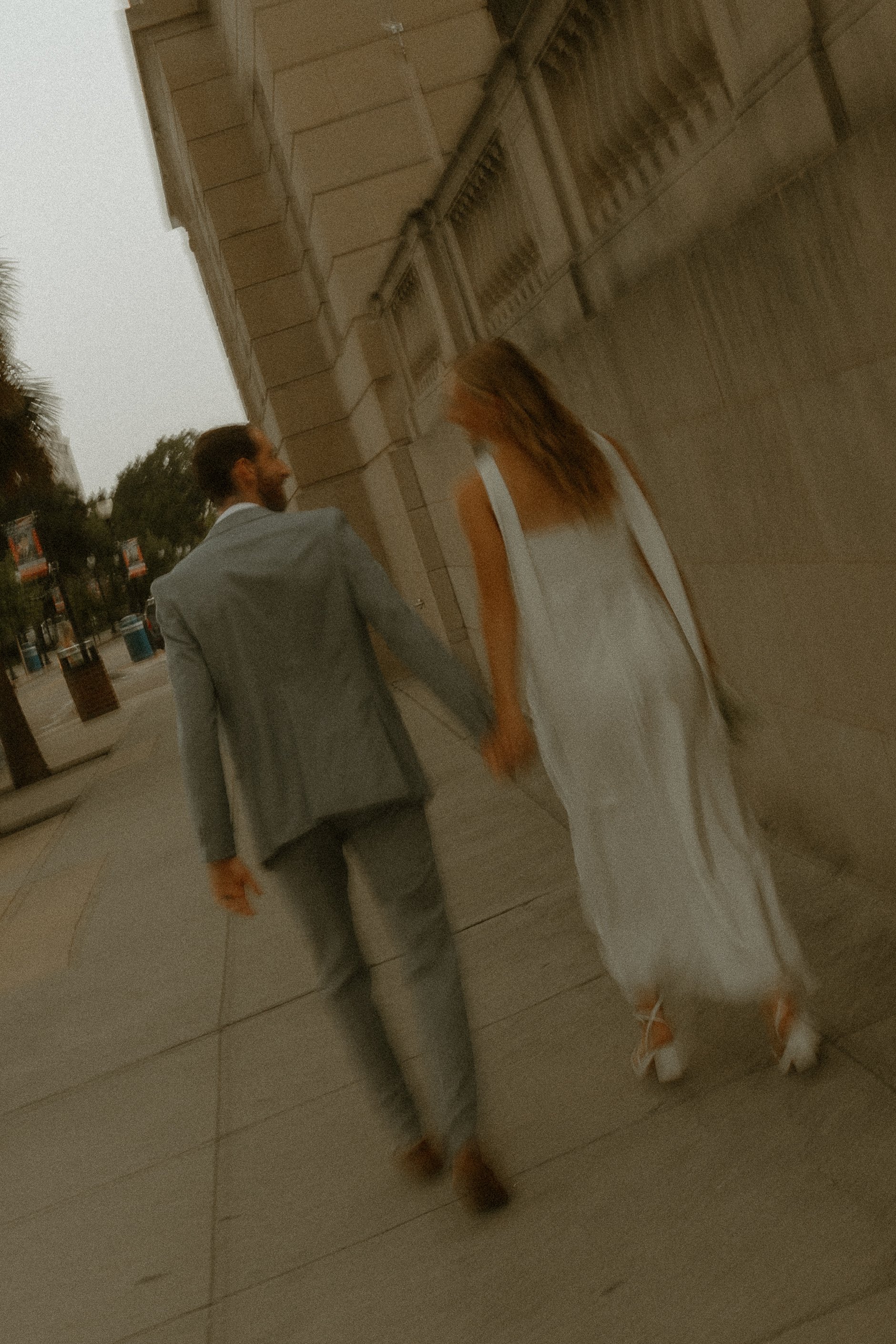 A man in a light gray suit and woman in a white dress walking together along a sidewalk next to a building with a metal fence, trees, and trash cans in the background.