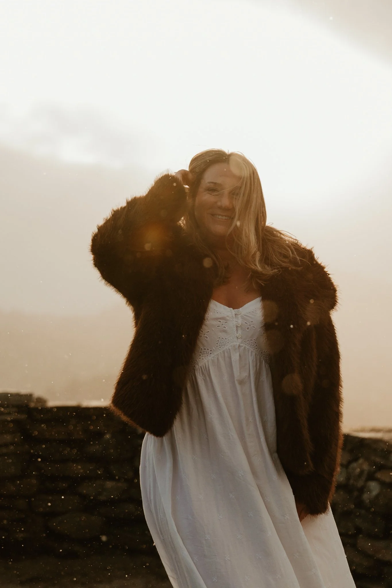 A woman smiling at the camera, wearing a white dress and a fur jacket, outdoors during sunset with a cloudy sky in the background.