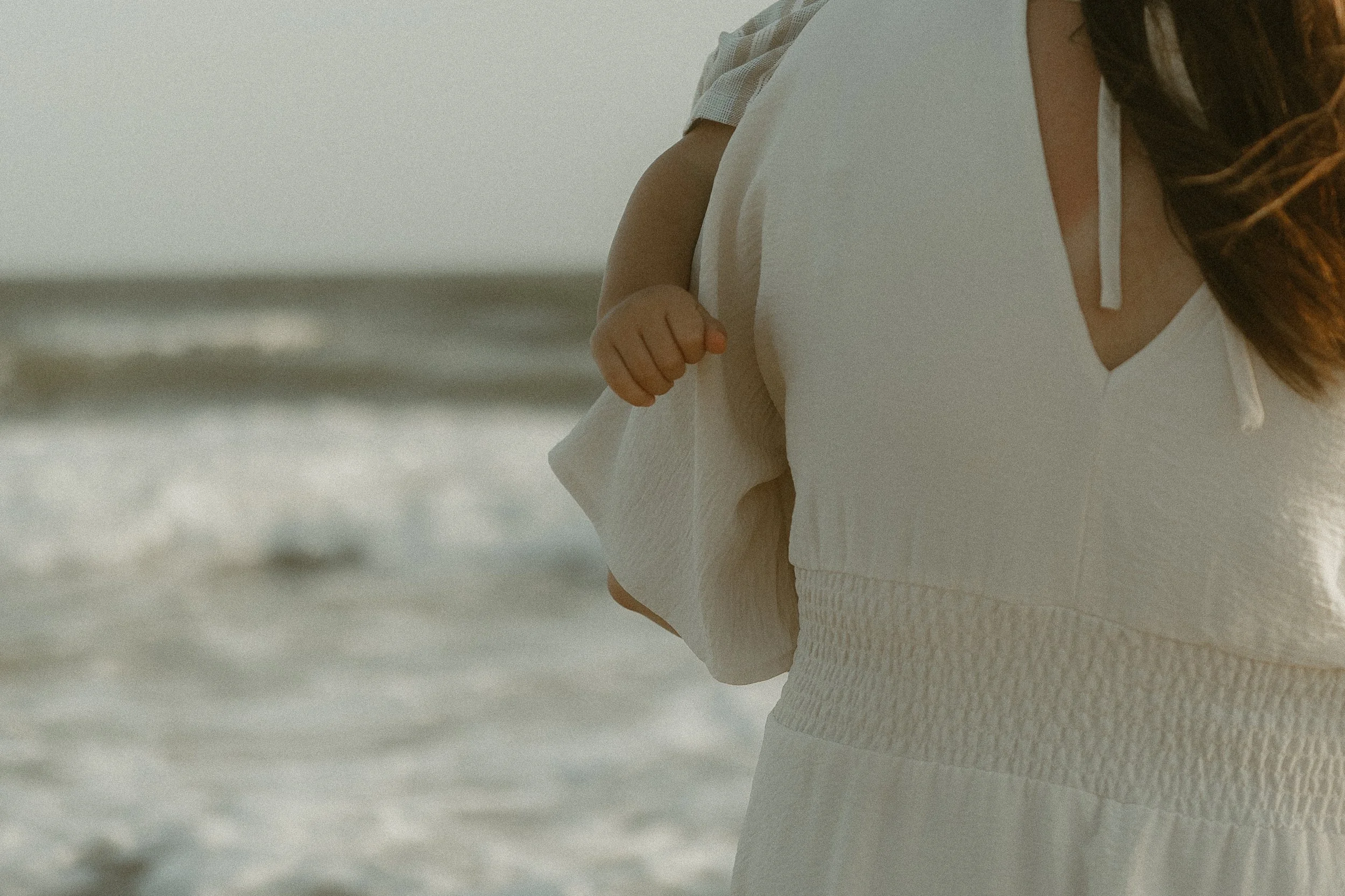 Close-up of a woman in white clothing walking on the beach, with a child's hand holding her dress.