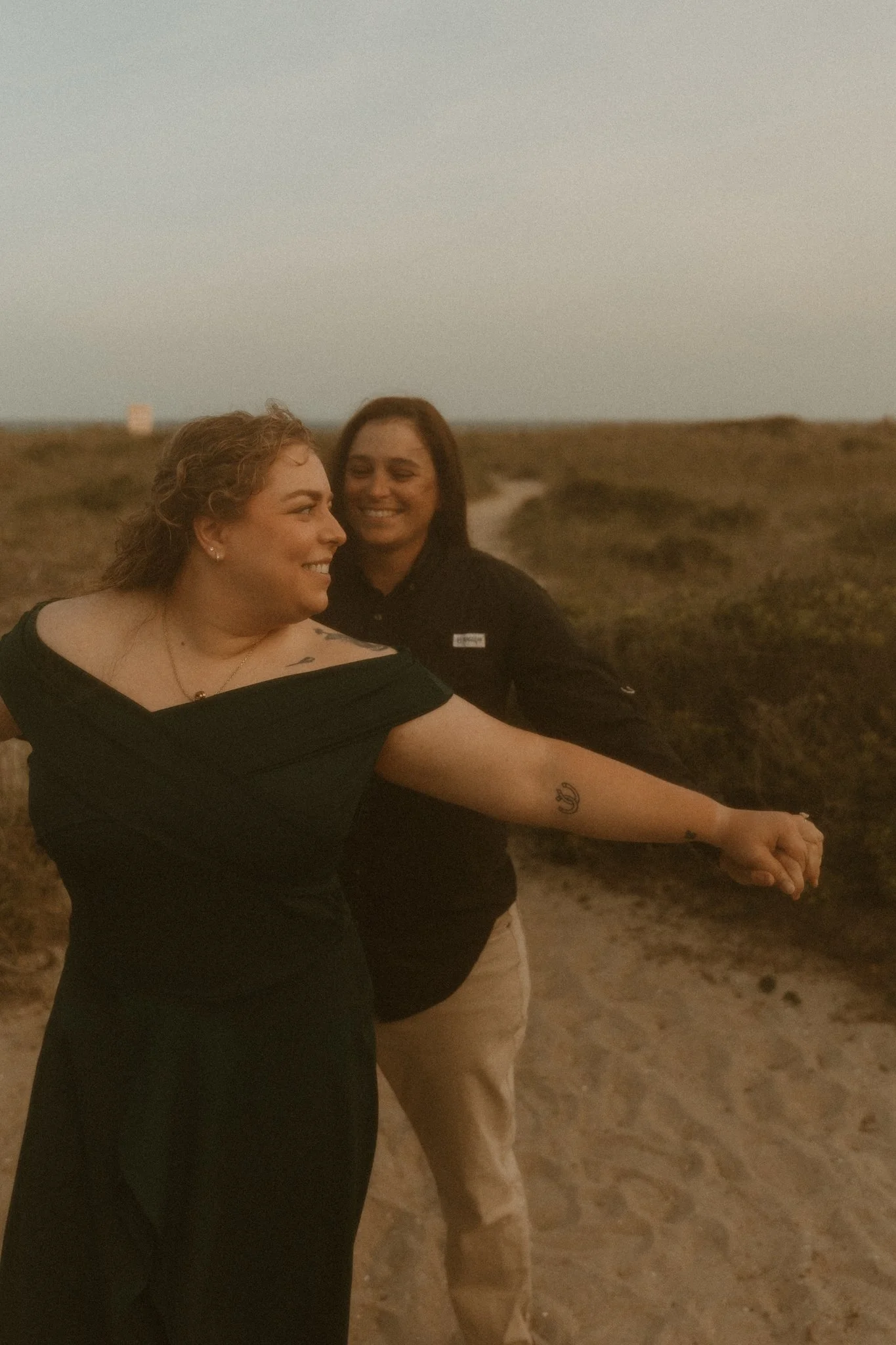 Two women standing on a sandy beach, one in the foreground in a black dress, the other in the background in a black shirt and beige pants, both smiling and looking at each other during sunset.