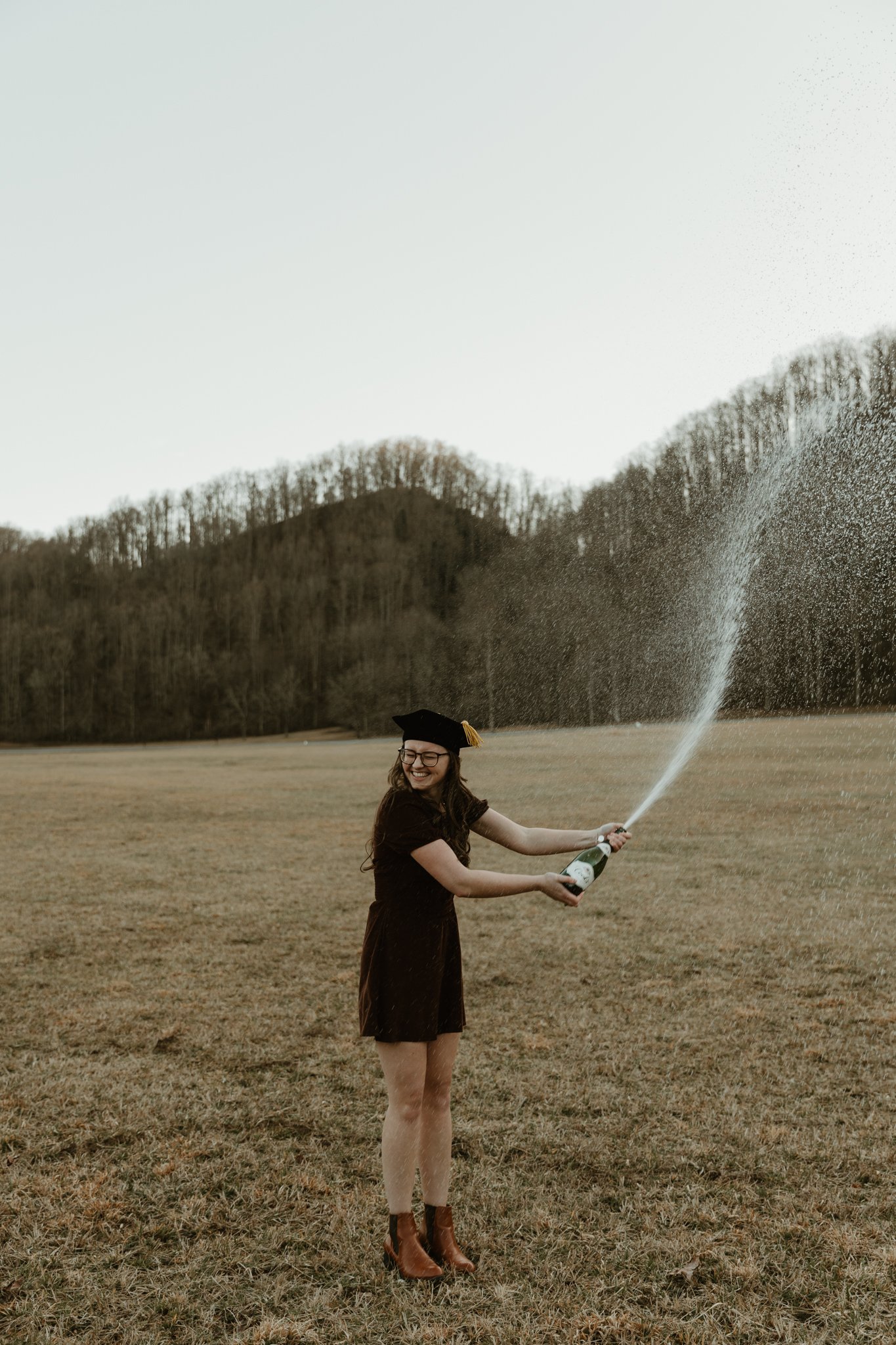 A woman wearing a graduation cap and dress celebrating outdoors by opening a bottle of champagne, with champagne spraying into the air, in a field with hills and trees in the background.