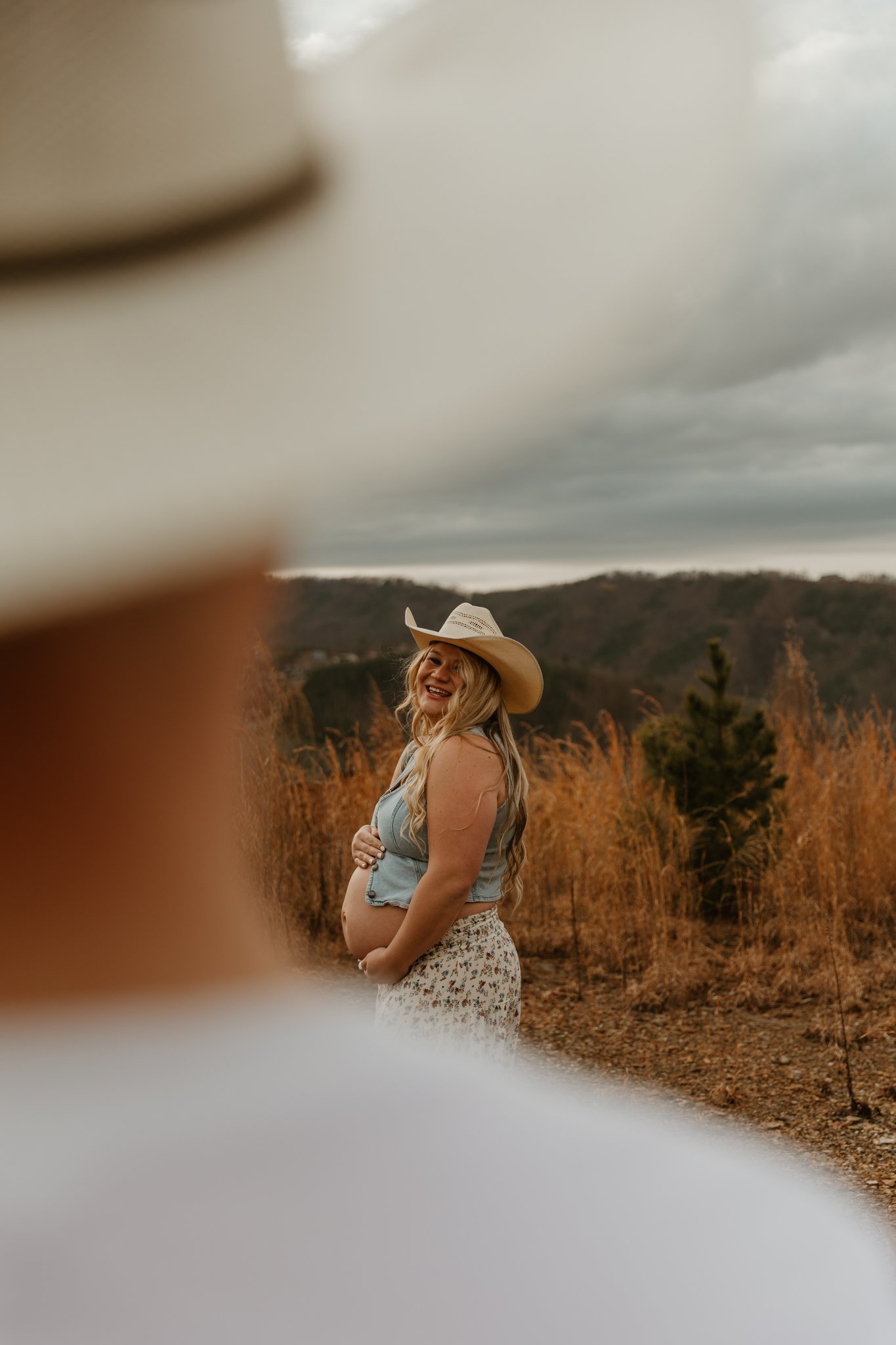 A pregnant woman smiling and posing outdoors in a field with tall grasses and mountains in the background, wearing a cowboy hat, sleeveless denim vest, and floral skirt.