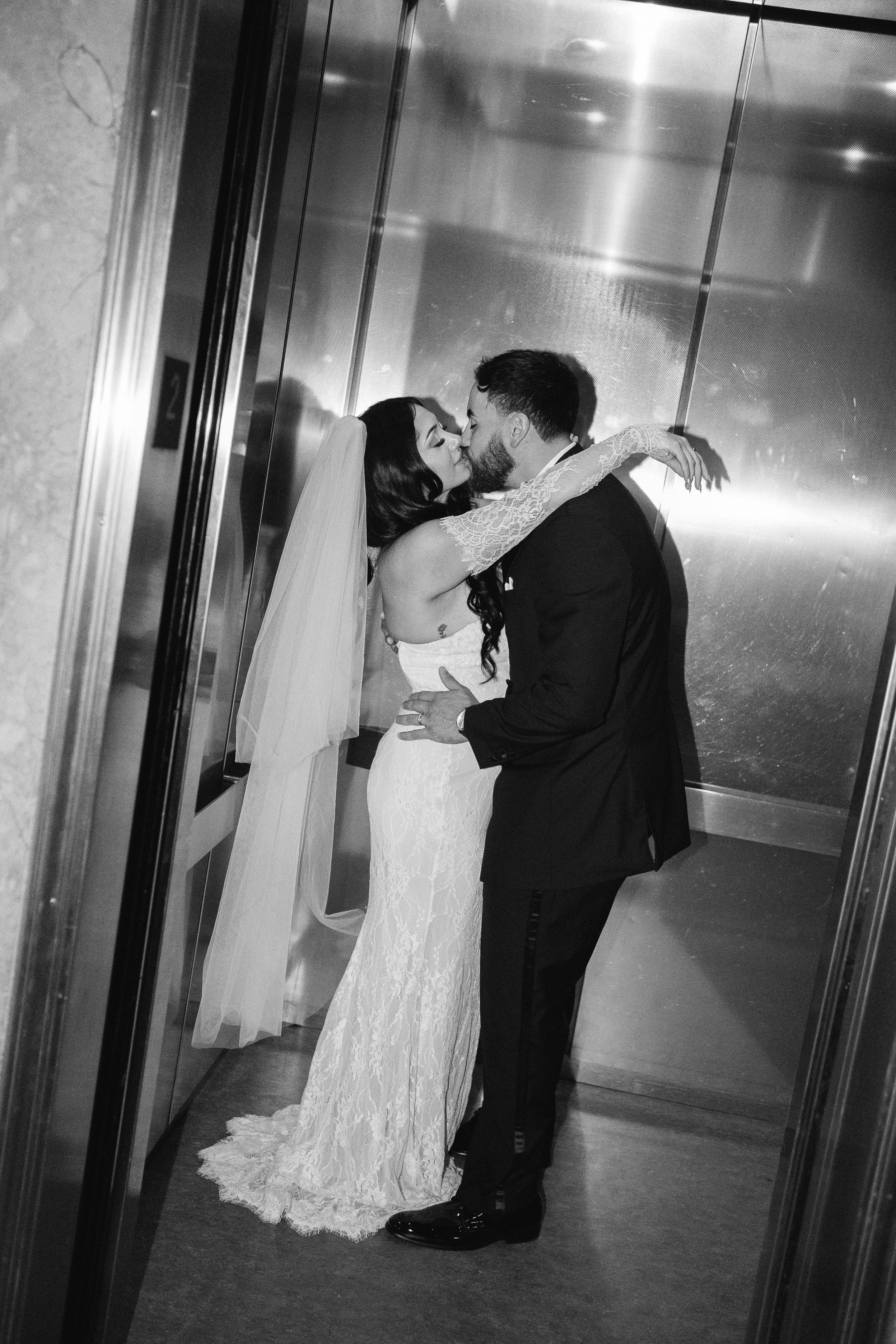 A couple in wedding attire sharing an intimate kiss in an elevator, with the bride wearing a lace wedding gown and veil, and the groom in a dark suit inside the Buncombe County Courthouse elopement in Asheville North Carolina