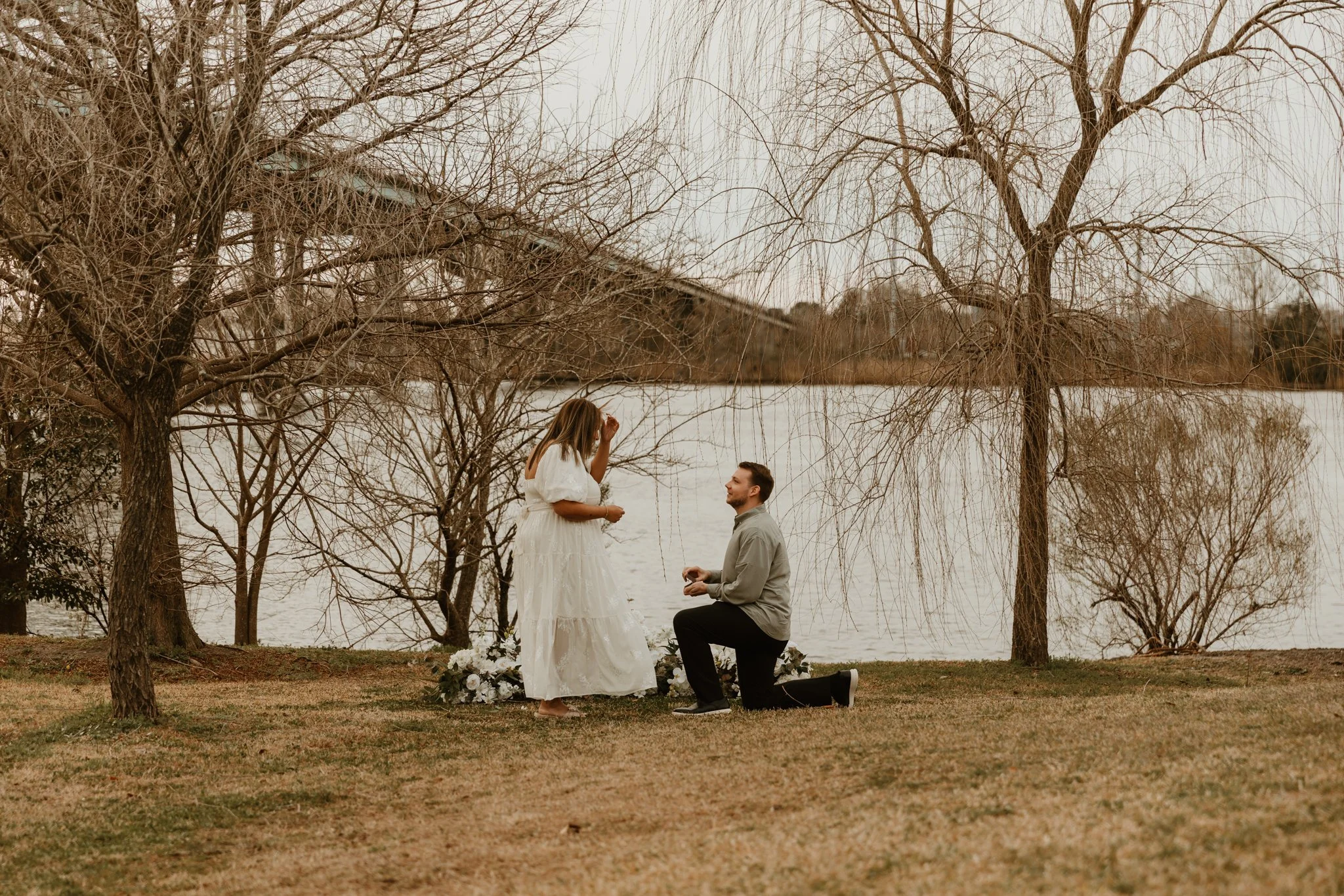 A couple is outside near a lake, with the man kneeling on one knee and the woman standing, her hand near her face, during a proposal or romantic moment on a grassy area with leafless trees.