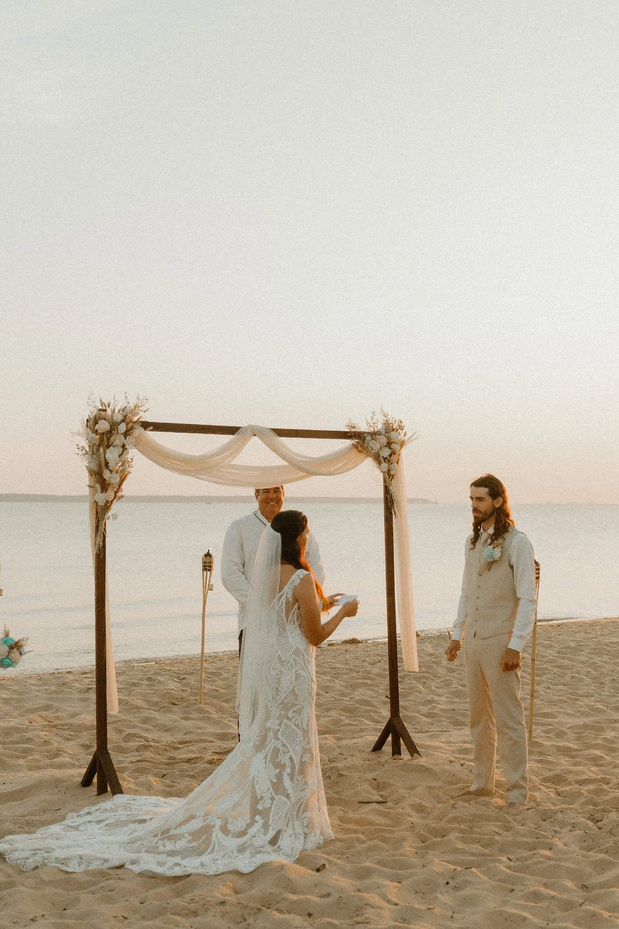 A couple gets married on the beach at sunset, with a wedding officiant and a bridesmaid in a white lace dress, under a decorated wooden arch with flowers.