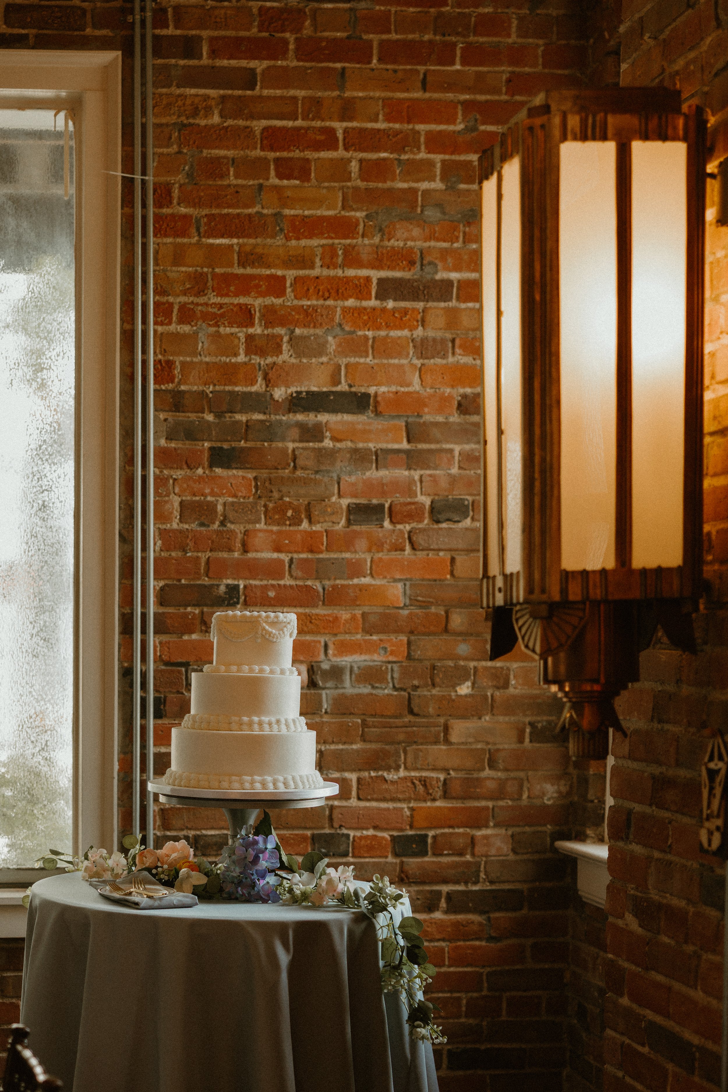 Elegant wedding cake with multiple tiers decorated with white icing, placed on a table with floral arrangements in front of a brick wall and a lit lamp.