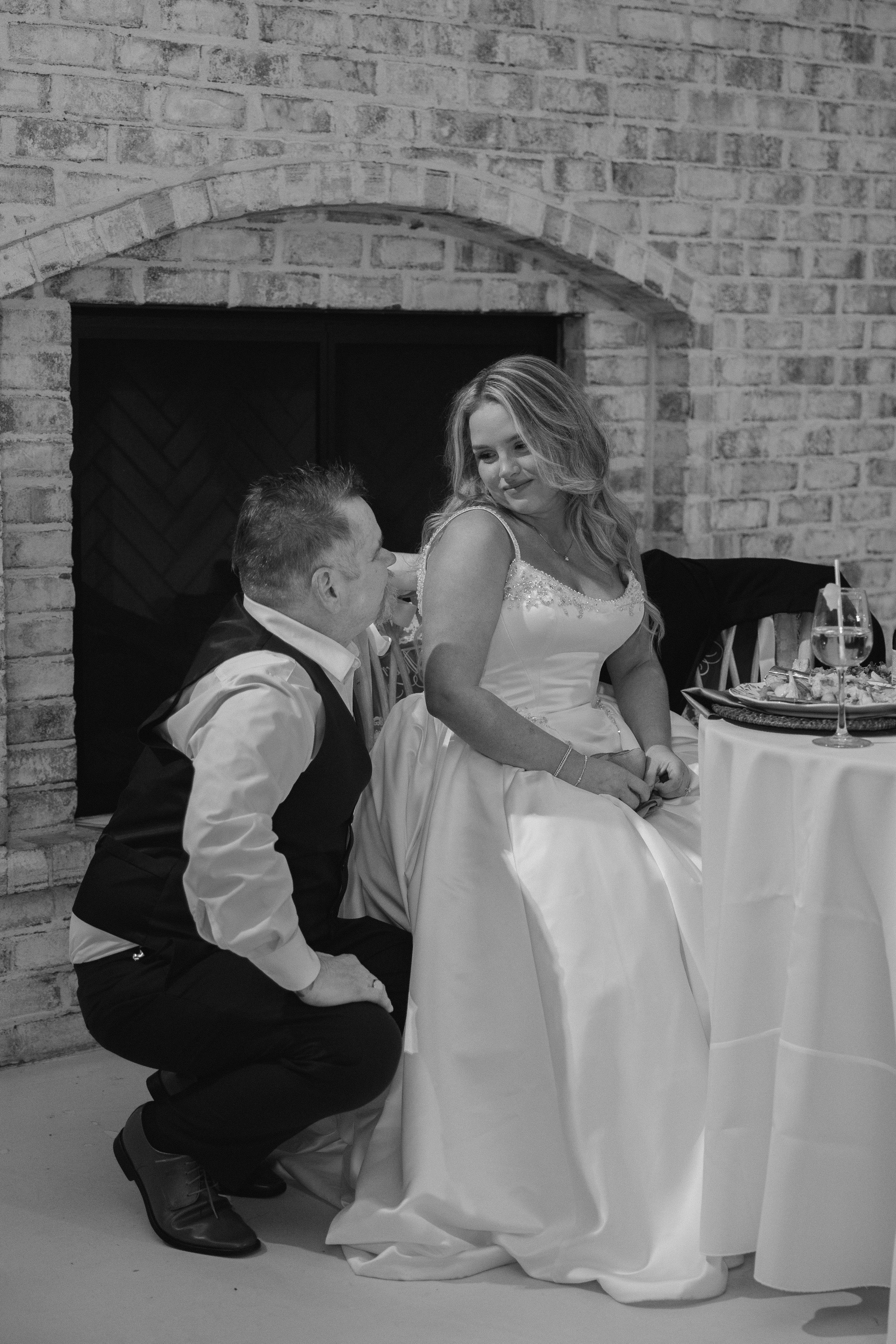 A black and white photo of a woman in a wedding dress sitting at a table, smiling and looking at a man in a vest and shirt who is kneeling in front of her. They are against a brick wall background, with a table set with drinks and plates nearby.