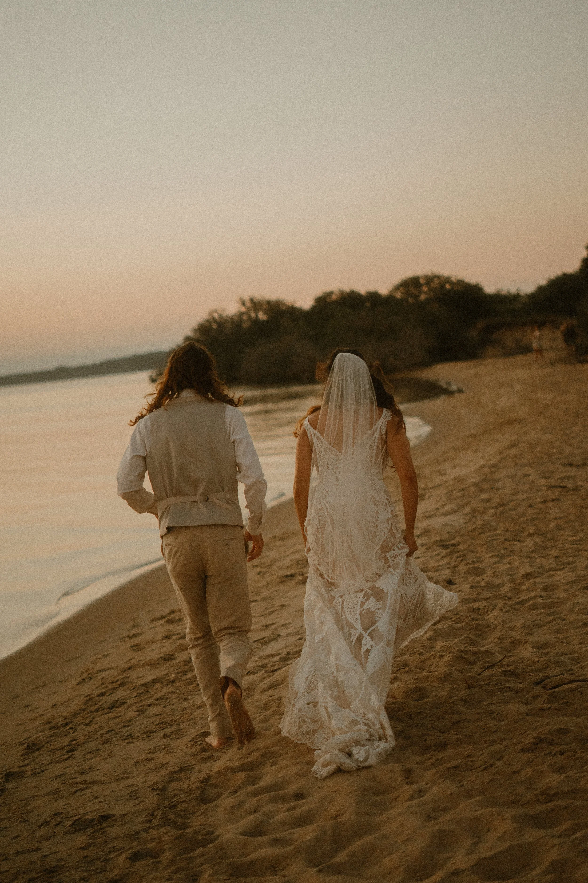 A couple dressed in wedding attire walking along a sandy beach during sunset, with water on the left and trees in the background.