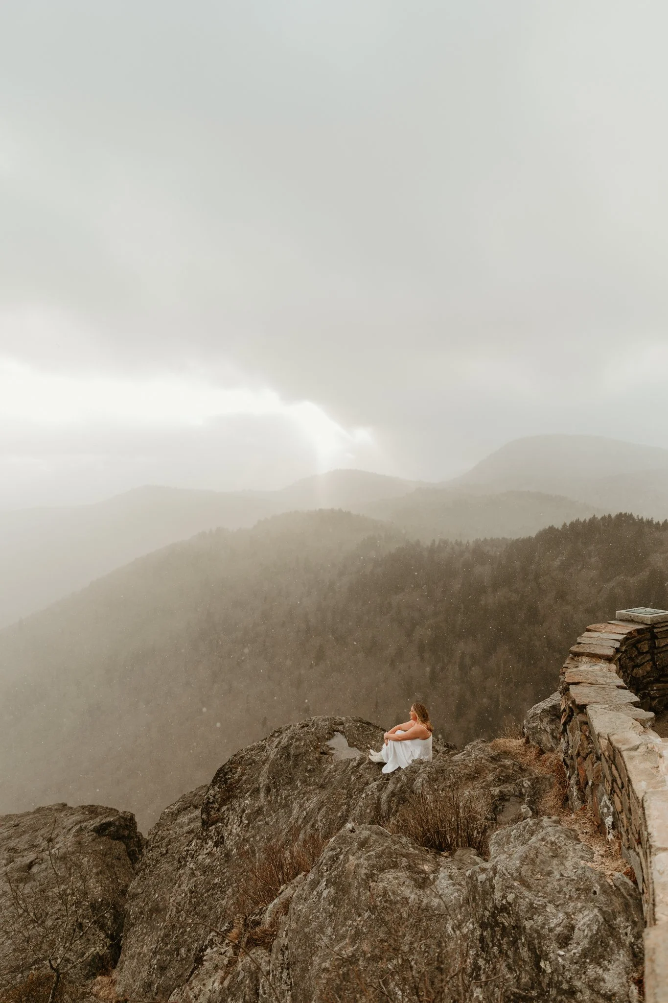 A woman in a white dress sitting on a rocky ledge overlooking a foggy mountain landscape with trees and distant hills.