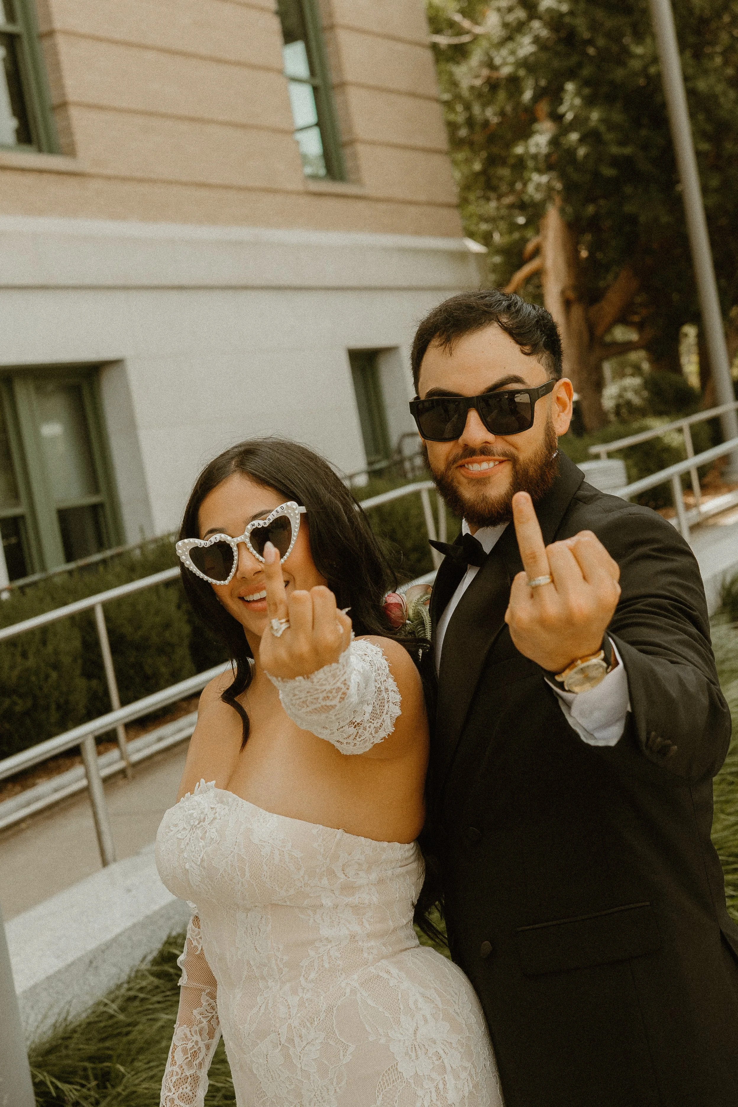 A newlywed couple in wedding attire making a gesture with their middle fingers raised, smiling for the camera outdoors near a modern building with large windows and trees in the background.