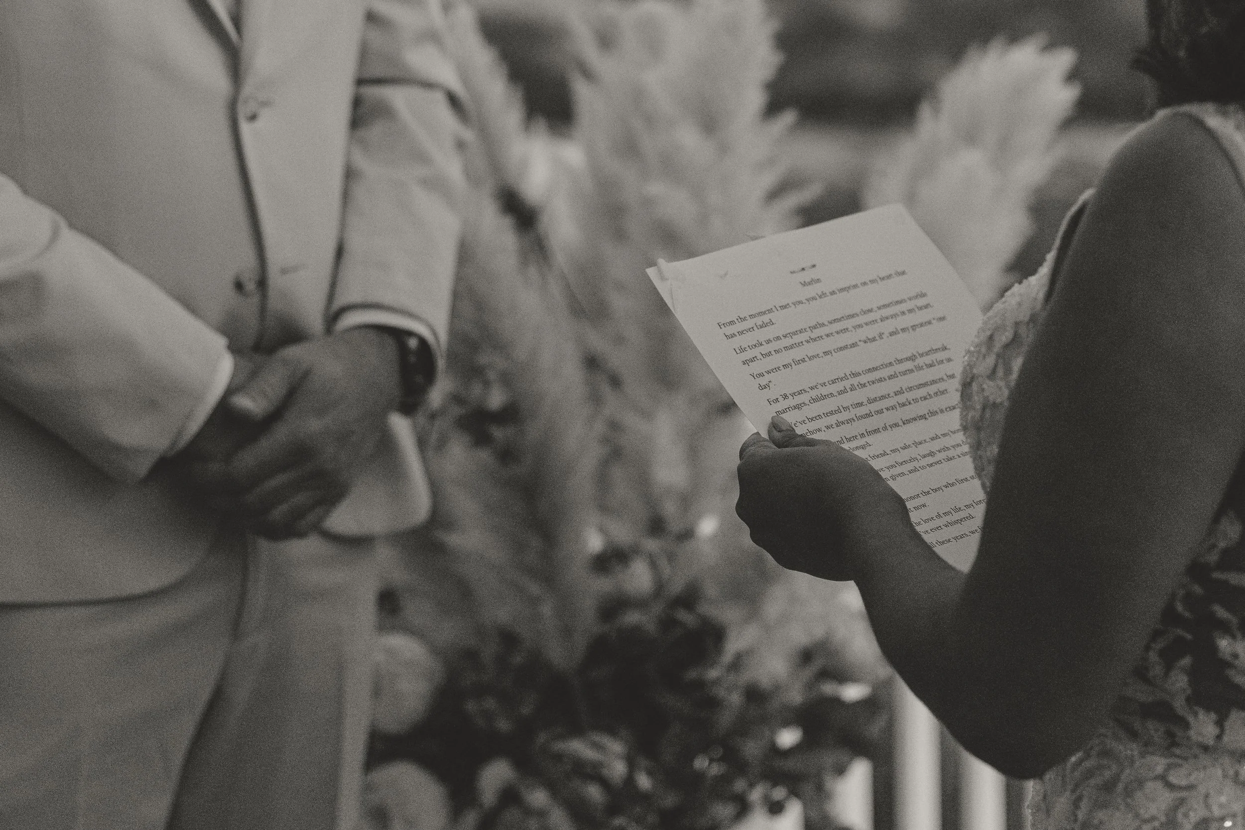 A black and white photo of two people holding hands, one reading a letter with a blurred background of a natural outdoor setting.