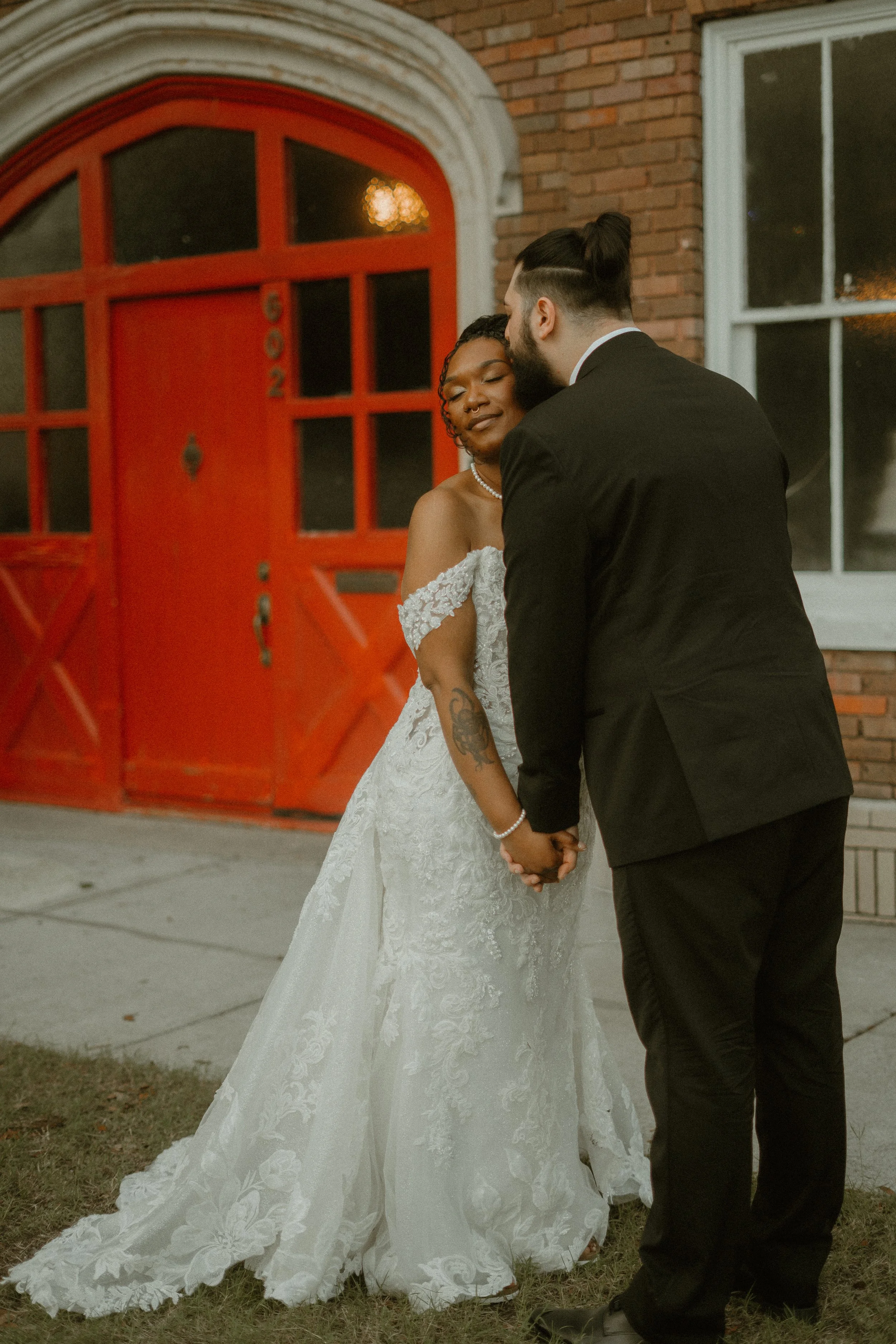 A bride and groom holding hands and kissing outside a brick building with a red arched door and a window.