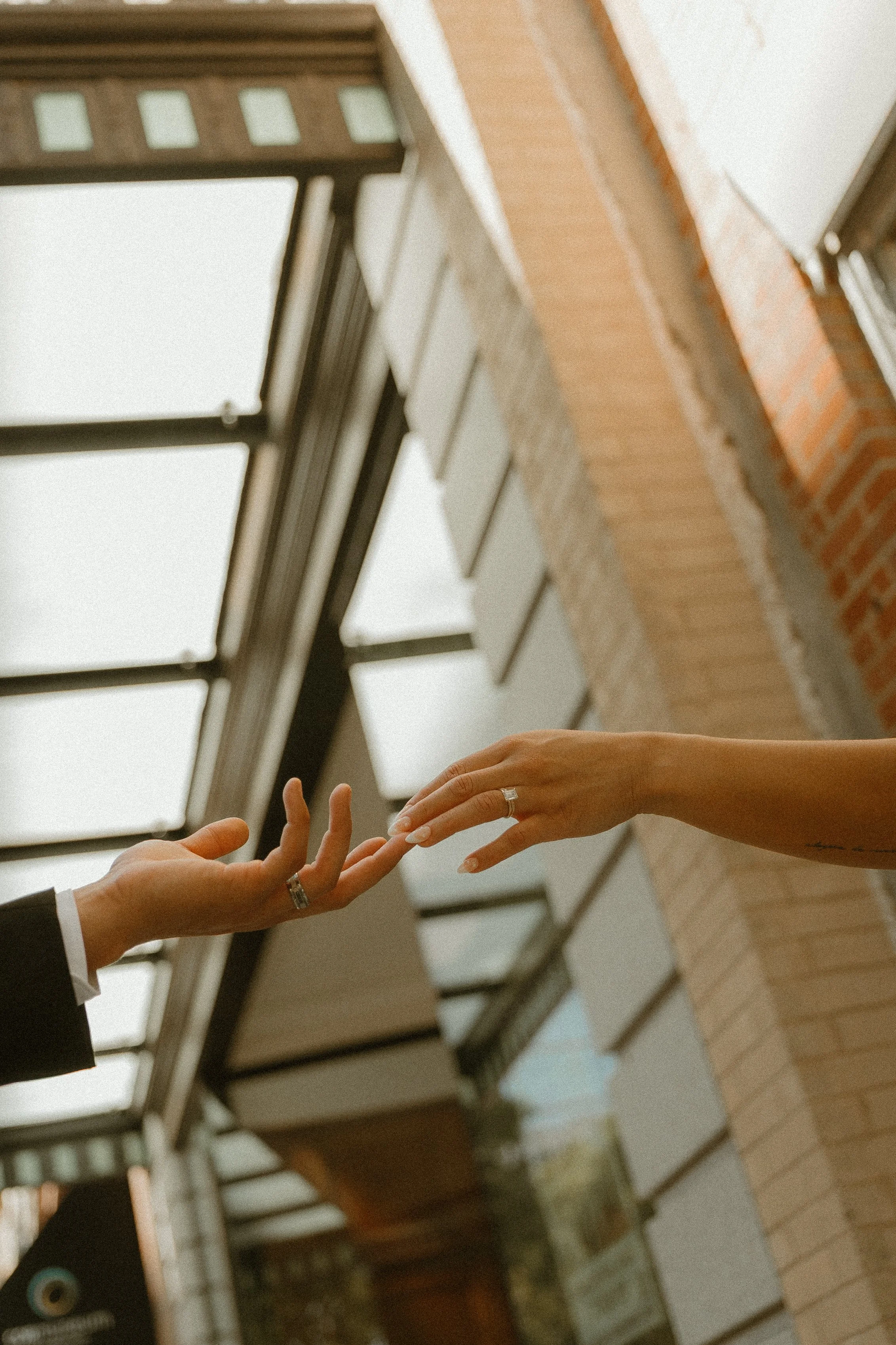 Two hands reaching towards each other, with one hand wearing a ring, in front of a brick building and large window.