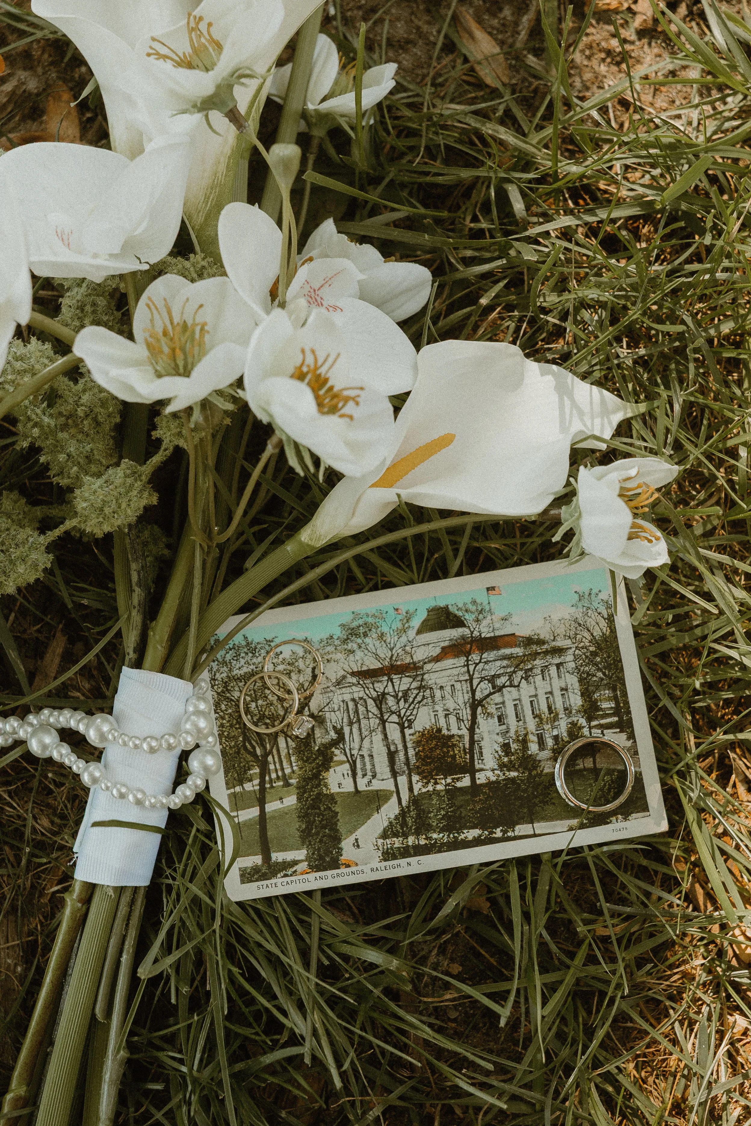 White flowers, pearl and gold jewelry, and an old photograph of the North Carolina State Capitol are lying on grass.